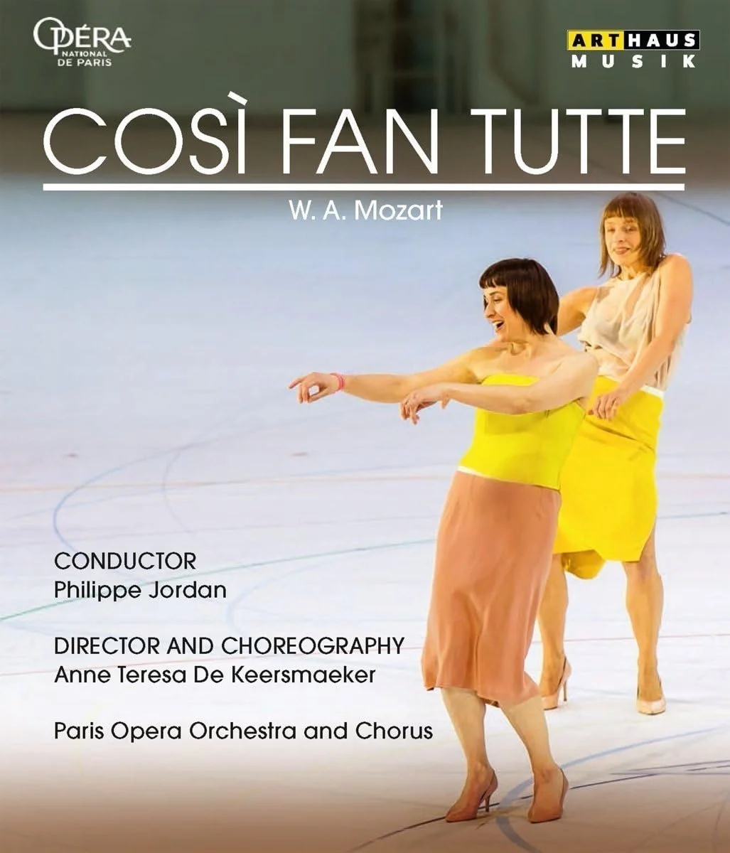 Poster for a performance of W. A. Mozart's 'Cosi Fan Tutte' featuring two women in yellow skirts and tops, one smiling and pointing, on an indoor stage with a blurred background.