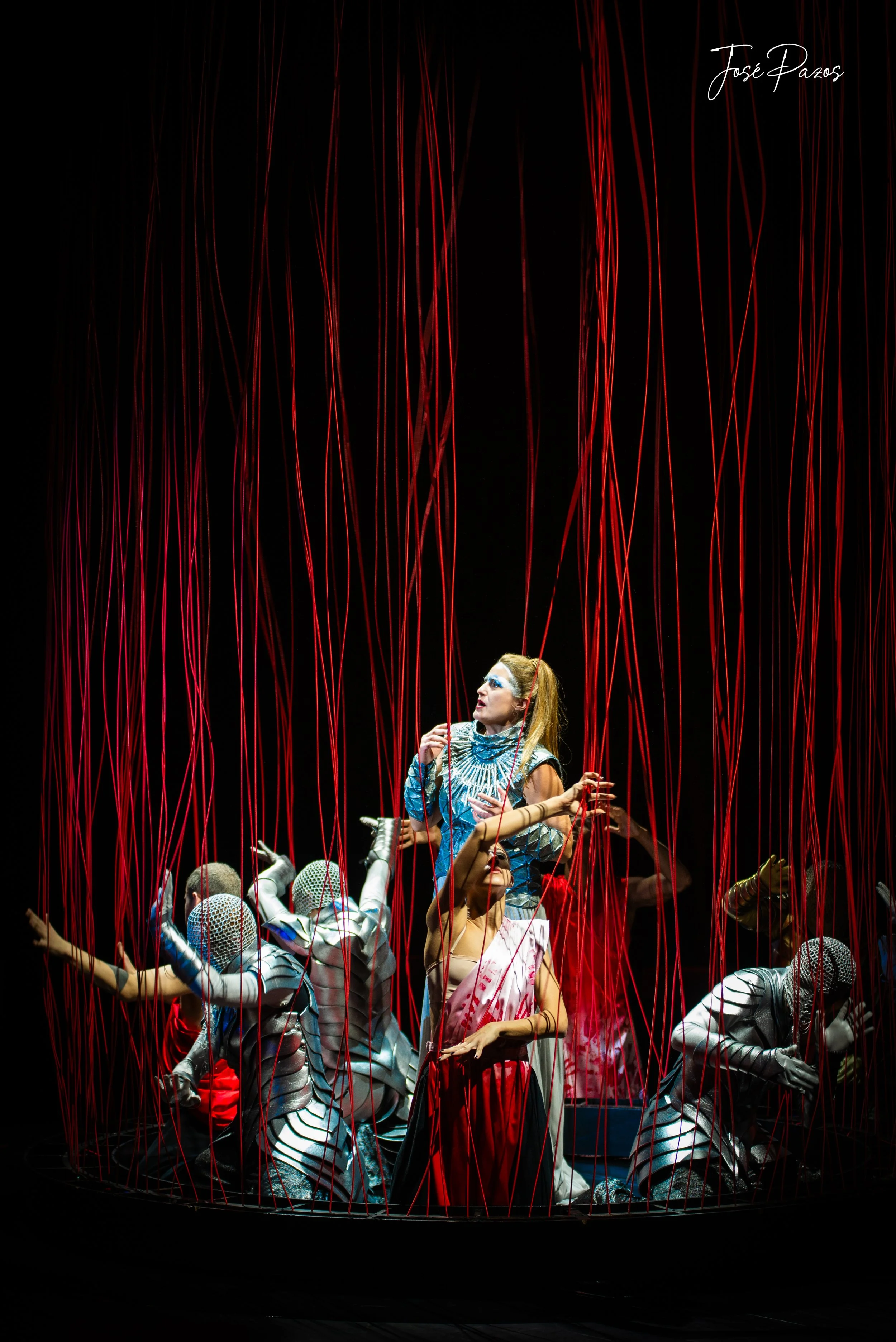 The image shows a theatrical performance with a woman dressed in a metallic costume and blue makeup, surrounded by performers in shiny silver outfits with striped patterns. Red ribbons hang above them, creating a cage-like environment.