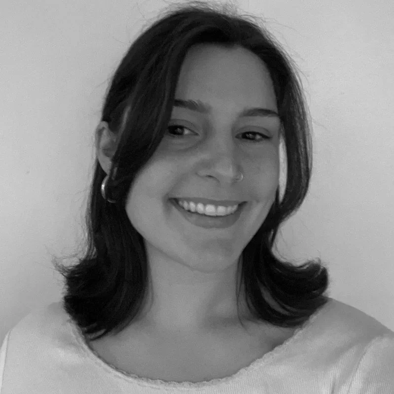 Black and white photo of a smiling woman with shoulder-length dark hair, hoop earrings, and a nose ring, wearing a light-colored top.
