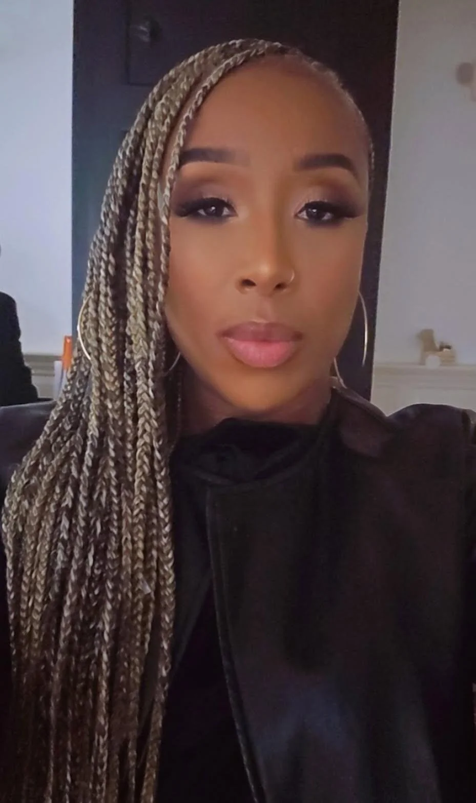 Close-up portrait of a woman with long braided hair, hoop earrings, and makeup, wearing a dark jacket, in an indoor setting.