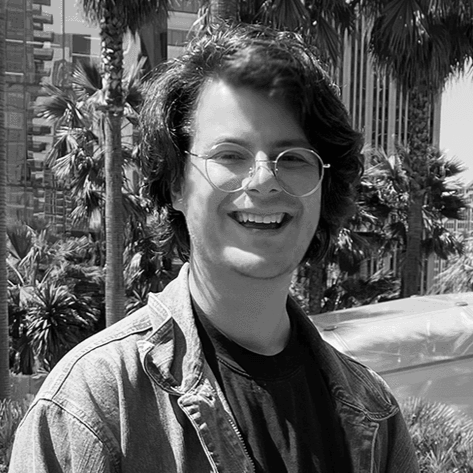 A smiling young man with glasses and wavy hair standing outdoors in front of trees and buildings, captured in black and white.