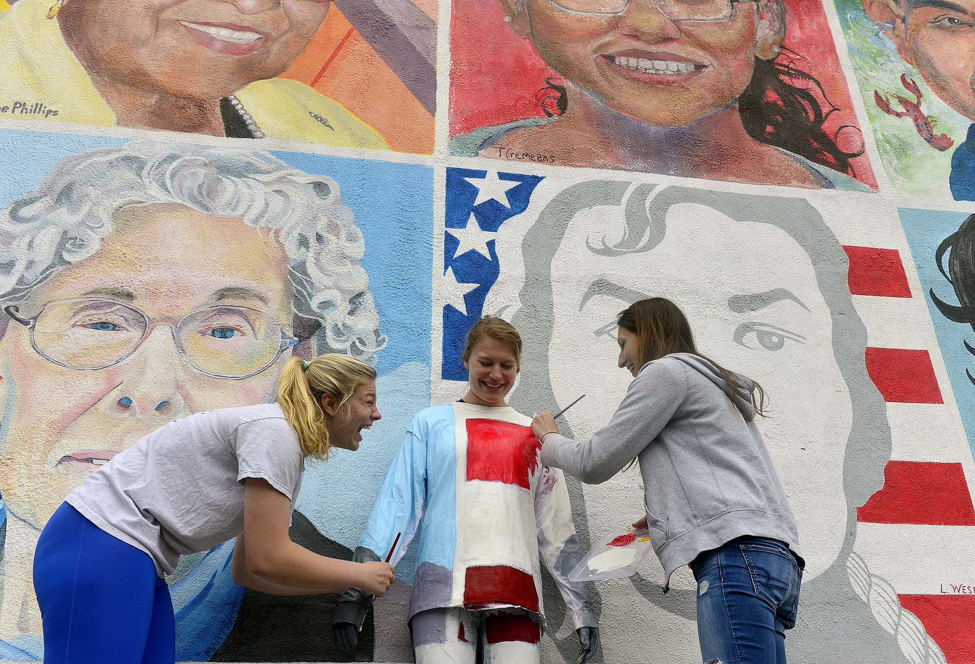 Connecticut College sophomores Emily Ehler, right, and Emily Senatore, left, laugh as they paint classmate Caitlyn Teare, center, into the "One Place Many Cultures" mural on Friday, May 12, 2017, as part of their final for a color theory class "Decoding Color" in downtown New London. For their final project class members worked in teams to find an urban space and camouflage a classmate into the environment. (The Day)