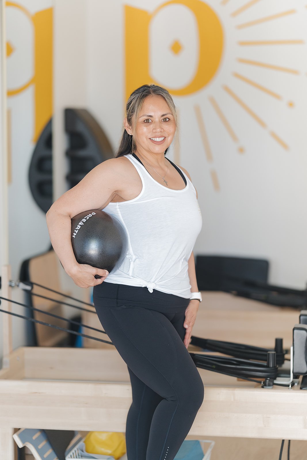 A woman in workout attire holding a black Medicine ball in her right arm at a pilates studio