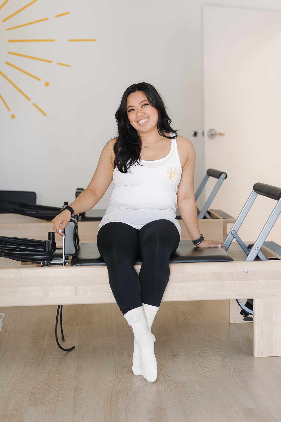 A woman sitting on a Pilates reformer machine, smiling, in a fitness studio with yellow wall art and light-colored wood flooring.