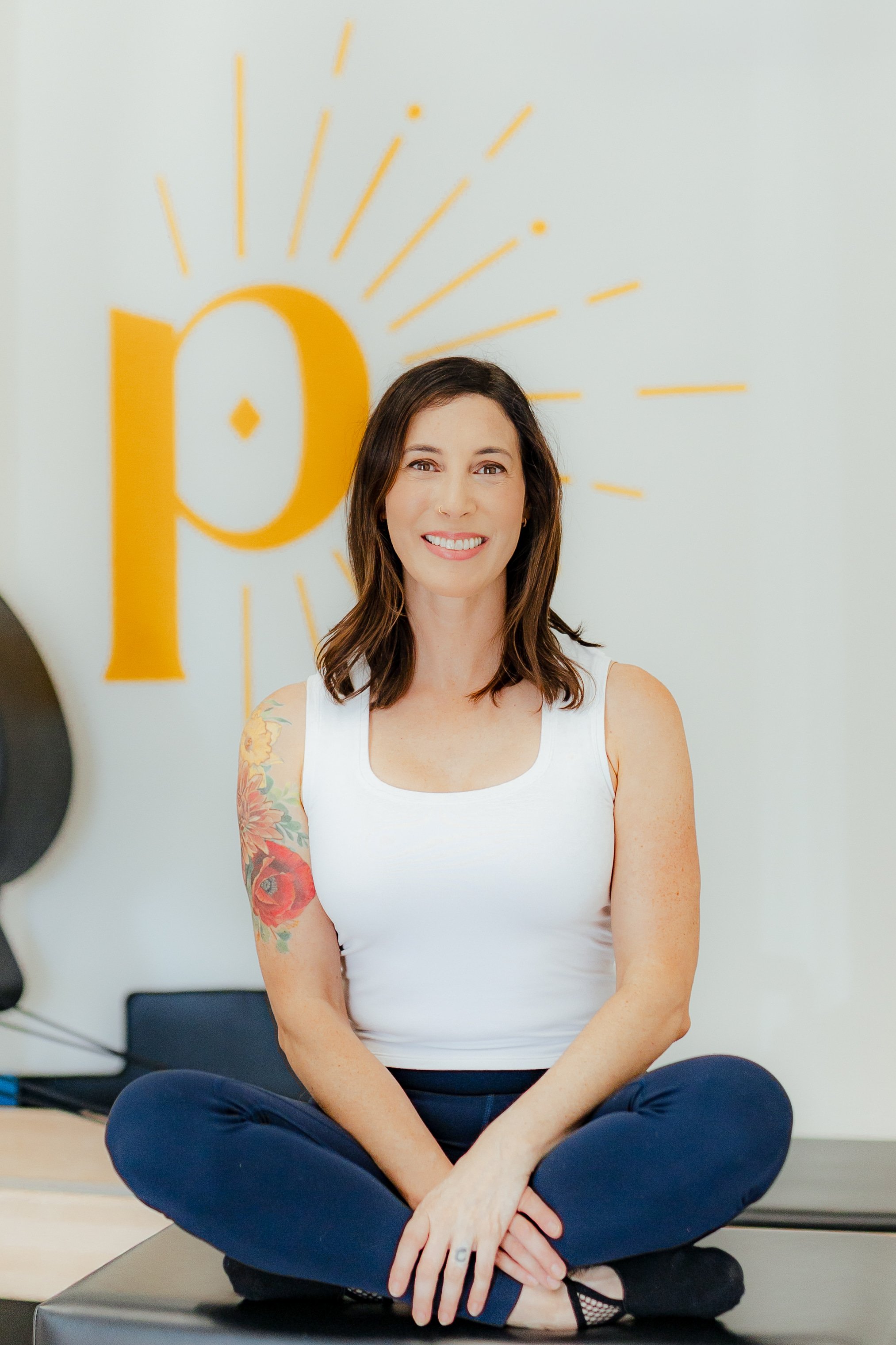 A woman sitting cross-legged on a black yoga mat, smiling, with a large yellow sun art in the background. She has shoulder-length brown hair, tattoos on her left arm, and wears a white sleeveless top and navy blue pants.