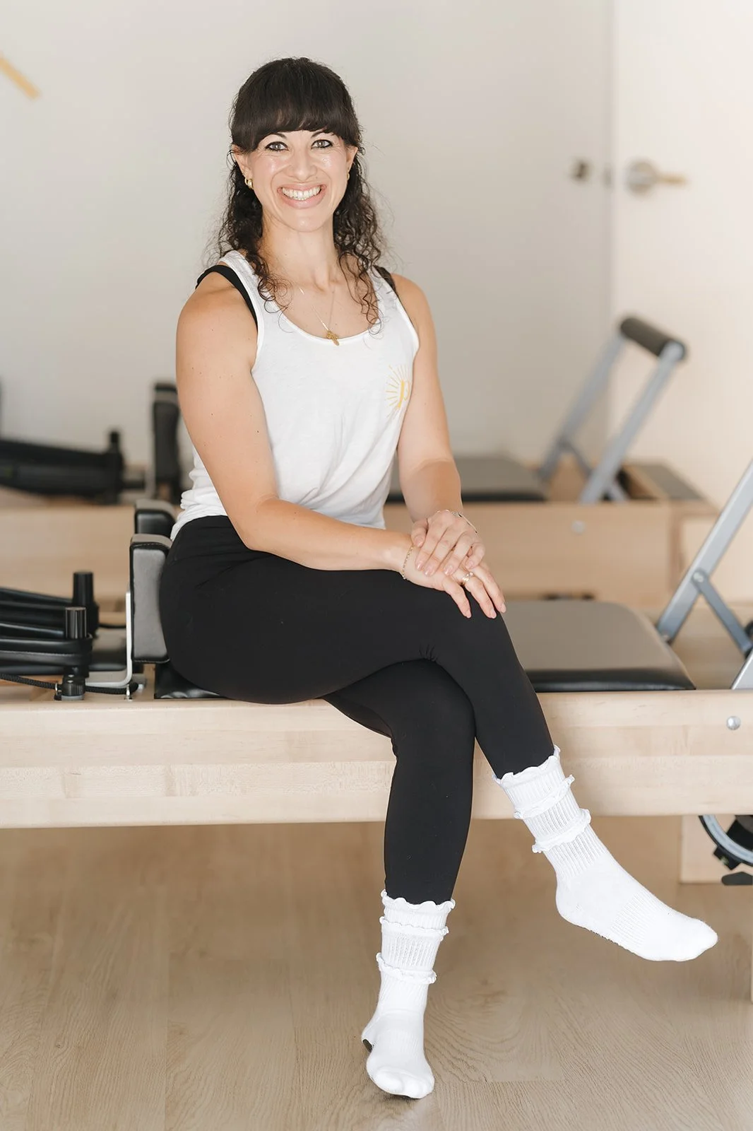 A woman with dark hair and bangs, smiling, sitting on a Pilates reformer machine in a pilates studio.