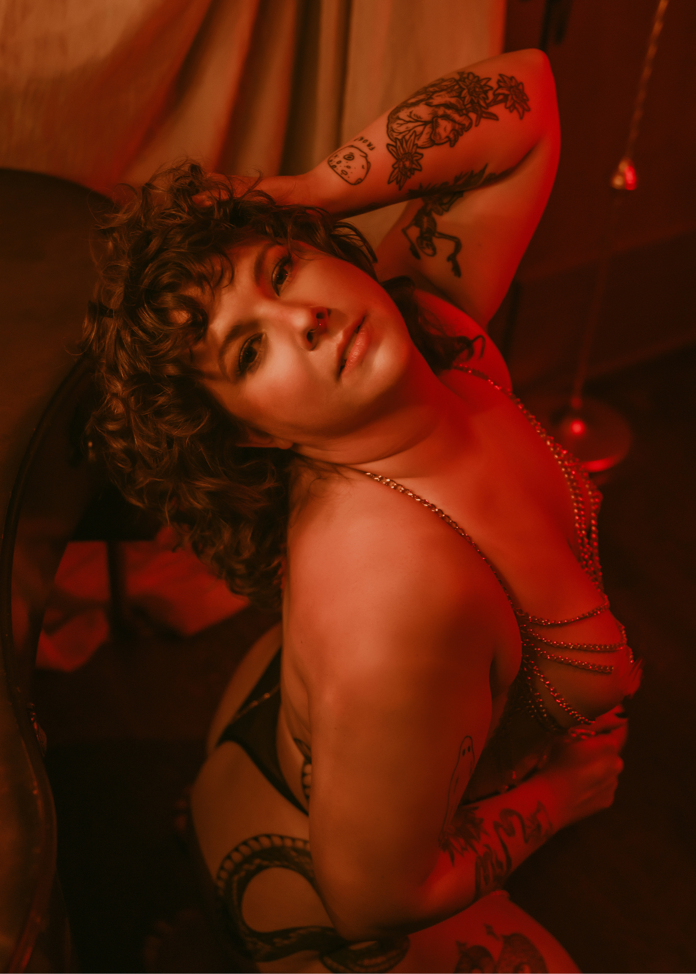 A woman with curly hair lying on a bed with red lighting, wearing black lingerie and displaying various tattoos.