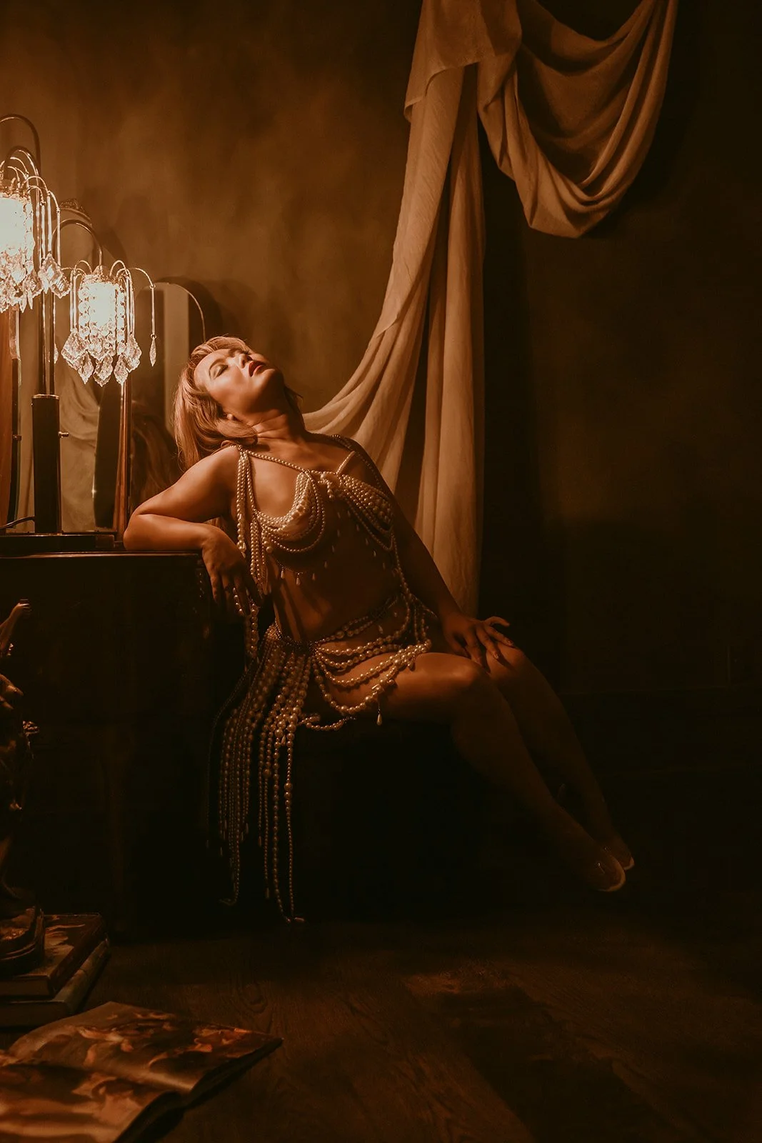 A woman posed on a dark bench or sofa, wearing an outfit made of pearl necklaces, in a dimly lit room with warm tones, curtains, and a vintage-style lamp.