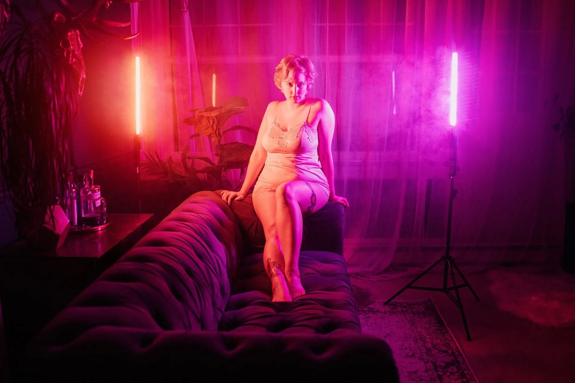 A woman in a beige slip dress sitting on a black velvet sofa in a room illuminated by pink and purple neon lights, with plants and a photography light stand in the background.