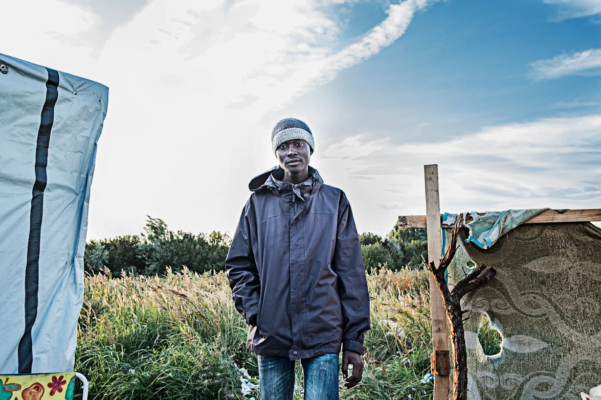 A young Sudanese man standing in the back of his tent where they have built an outdoor kitchen with his friends. Gave up to try to go to England as it is too risky, he applied for Asylum in France, having to wait in the jungle until he receives an answer about his case.
