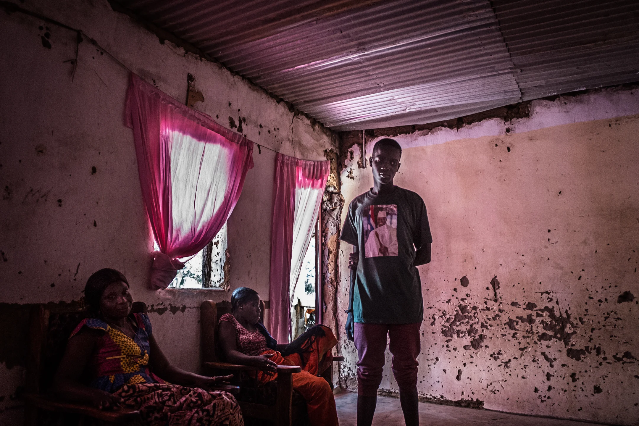 A young man standing in his family's living room wearing a t-shirt in support of former president Yahya Jammeh. When asked why he support Jammeh, the young man answer's " I just like his heart."