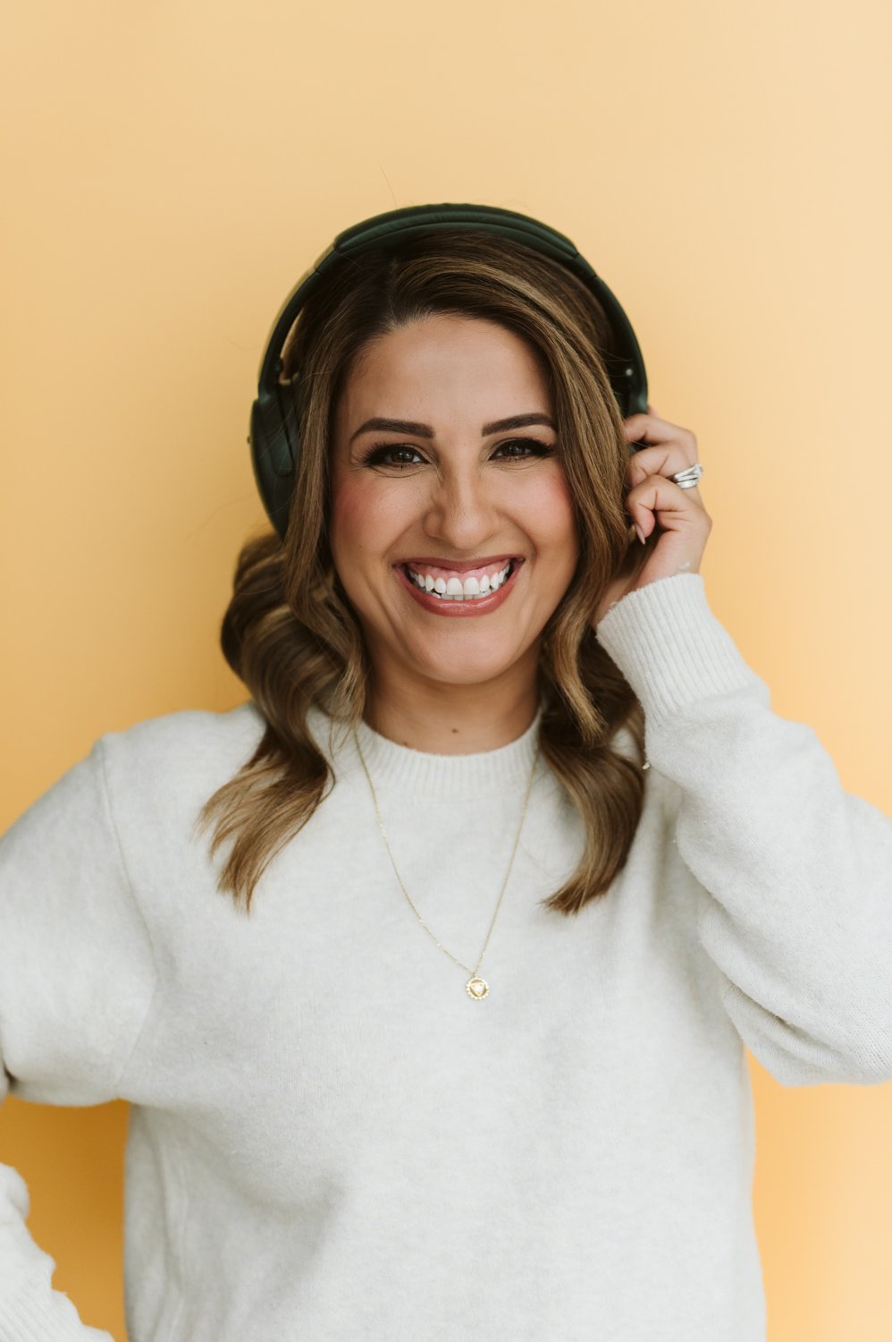 Personal branding photo of podcast host standing against yellow background, holding headphones and smiling