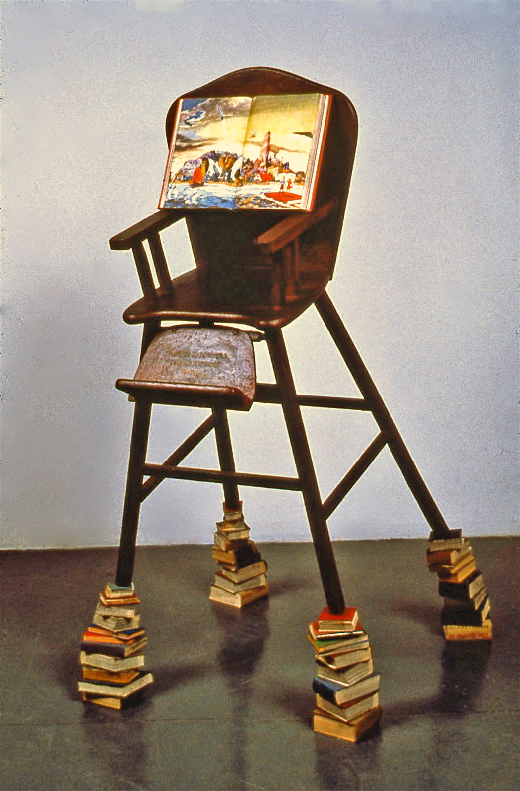 Harboring Illusions in the Sea of Information, 1989: chair, books, encaustic. Text on back of chair reads:
T he air is as warm as opium; the light diffuse, perfect, flattering. Small ships drift on the glassy water, their masts listing like happy drunks, while tiny whitecaps break on the shore with tactful grace. This could be home.
Animals walk towards us as we make for land. Their calm dark eyes are sympathetic and welcoming. They offer to carry your bags and cameras up the chalky cliffs.
High on the fissured slope, one stops to scratch a message, with stone, on stone: “Art is immortal, so are these words.” As he writes, it begins to rain, and the inscription melts away. Below us, in the water, the boats capsize. There is no going back. 