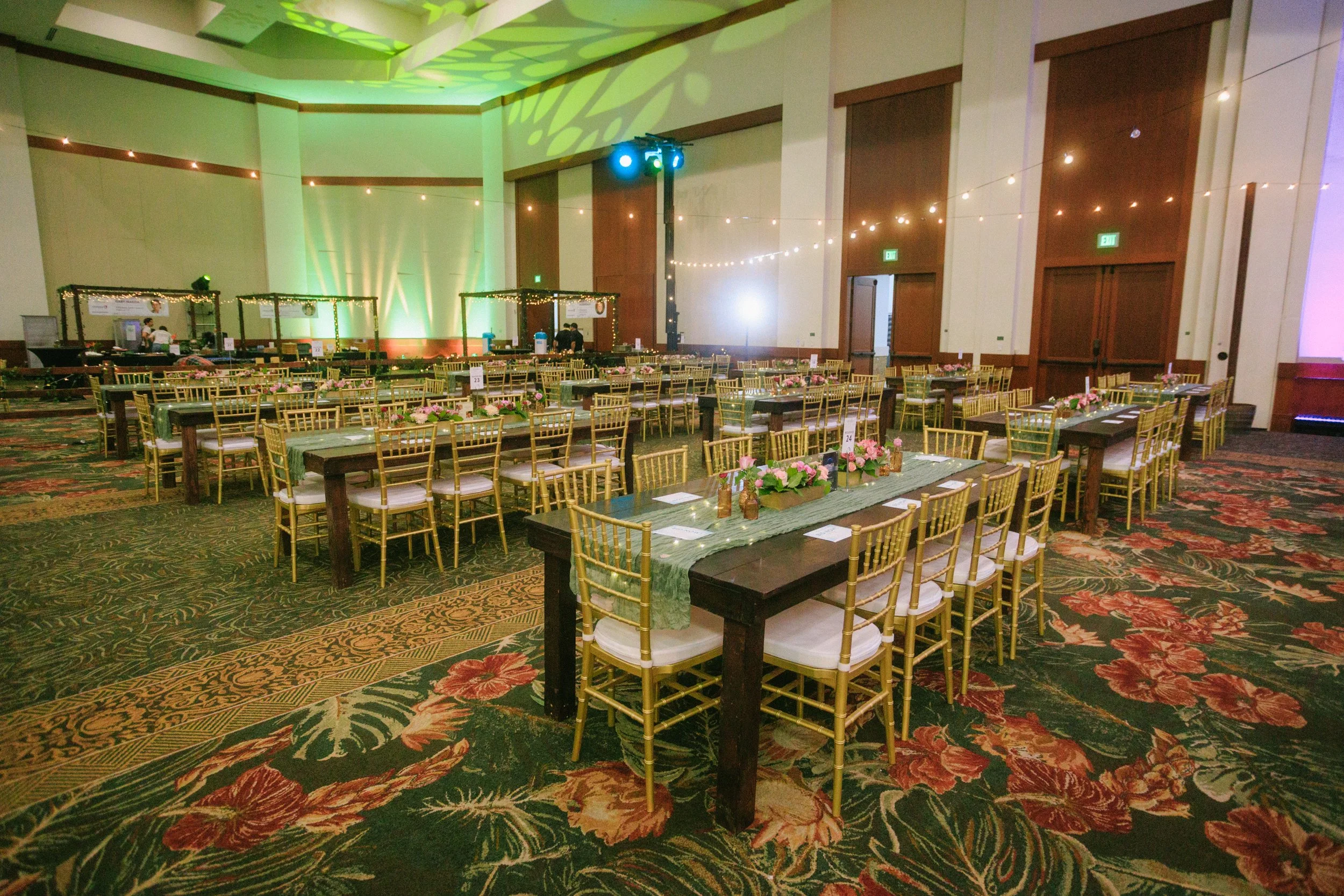 A banquet hall decorated for a celebration with long tables adorned with pink floral arrangements, gold chairs, and string lights on the walls. There are green uplights illuminating the background and a small stage at the front.