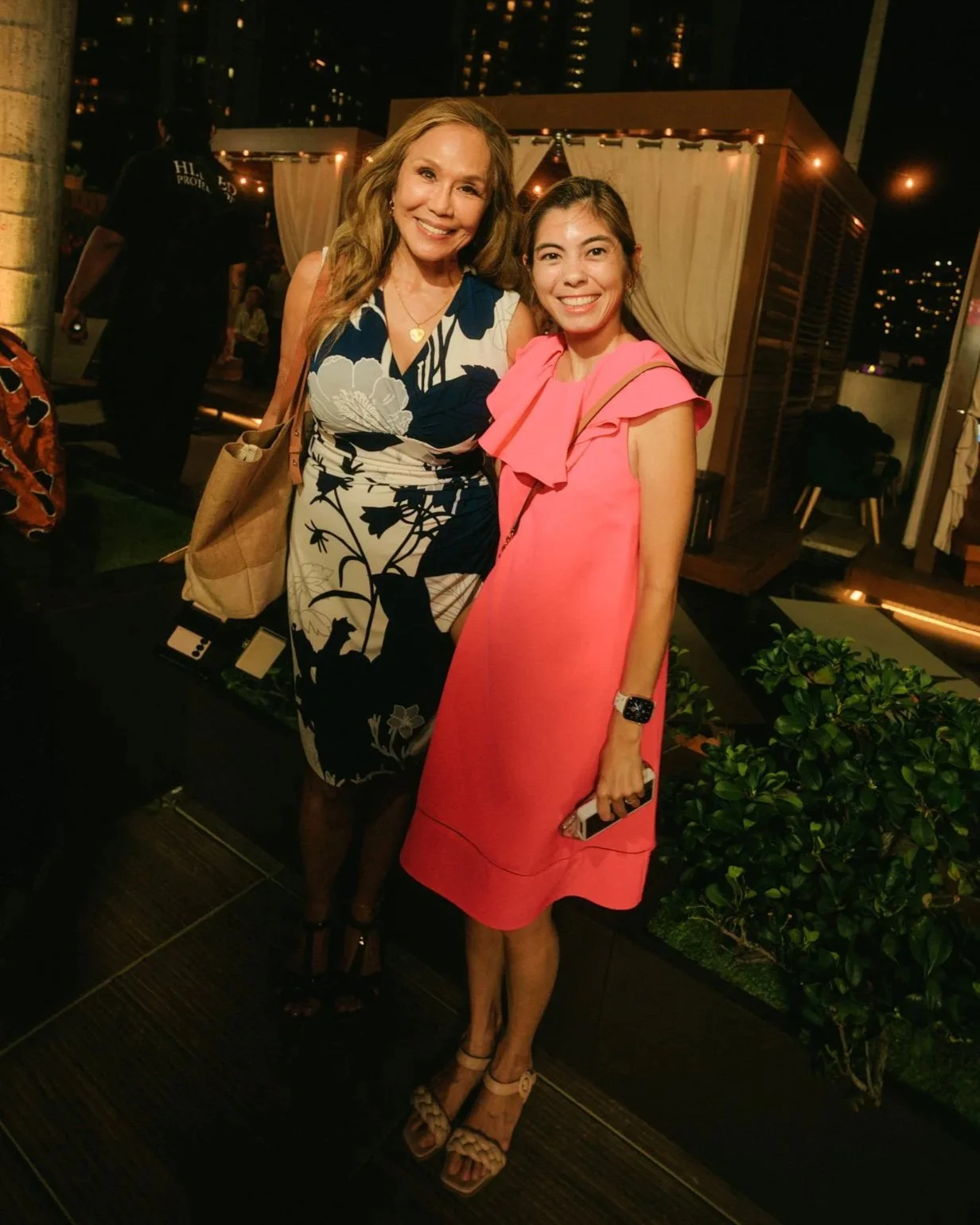 Two women standing close together and smiling at night. One in a dark floral dress, the other in a pink dress. There are string lights and a small outdoor setup in the background.