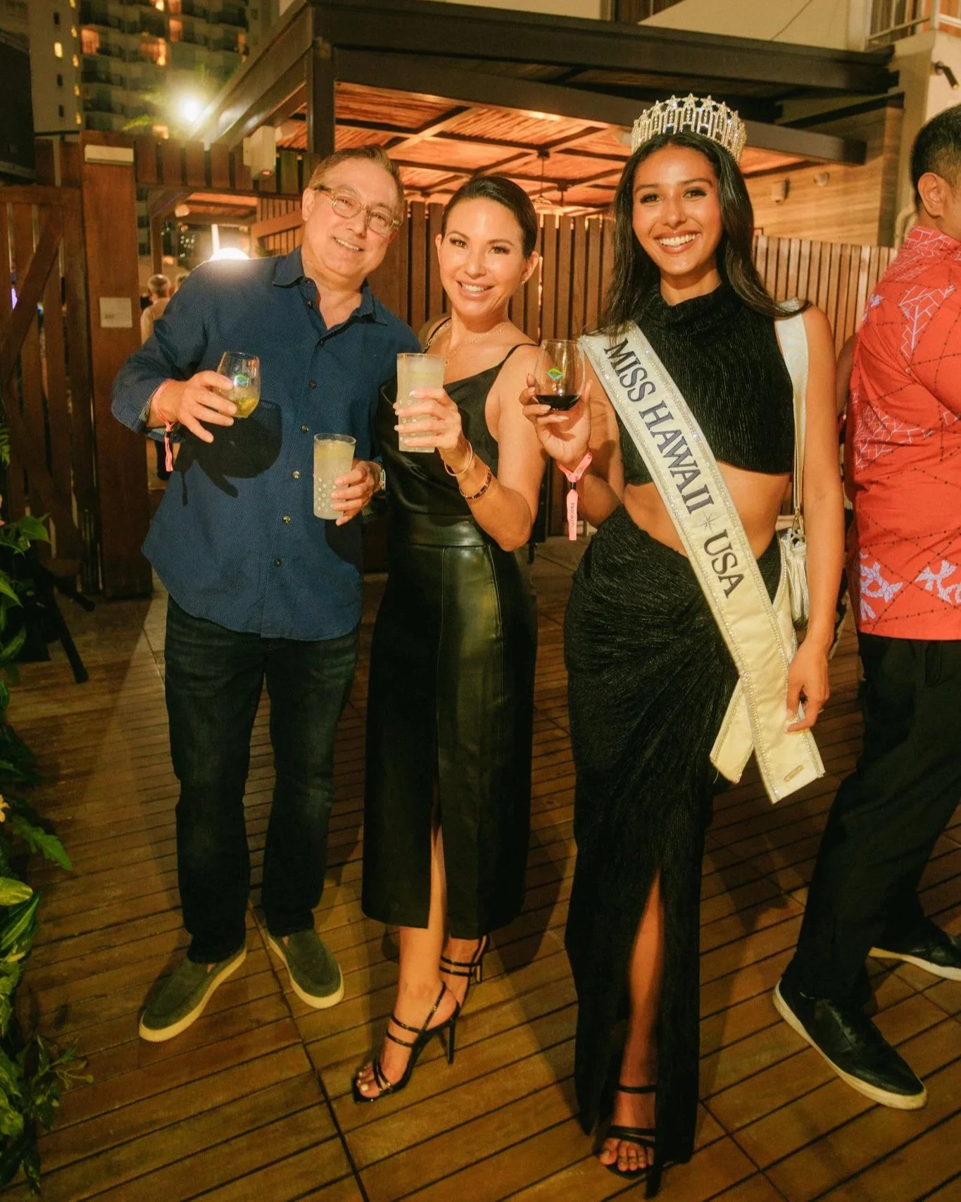 Three people at a celebration on a wooden deck, with a woman wearing a sash and crown holding a glass of red wine, flanked by two others holding drinks, smiling at the camera.