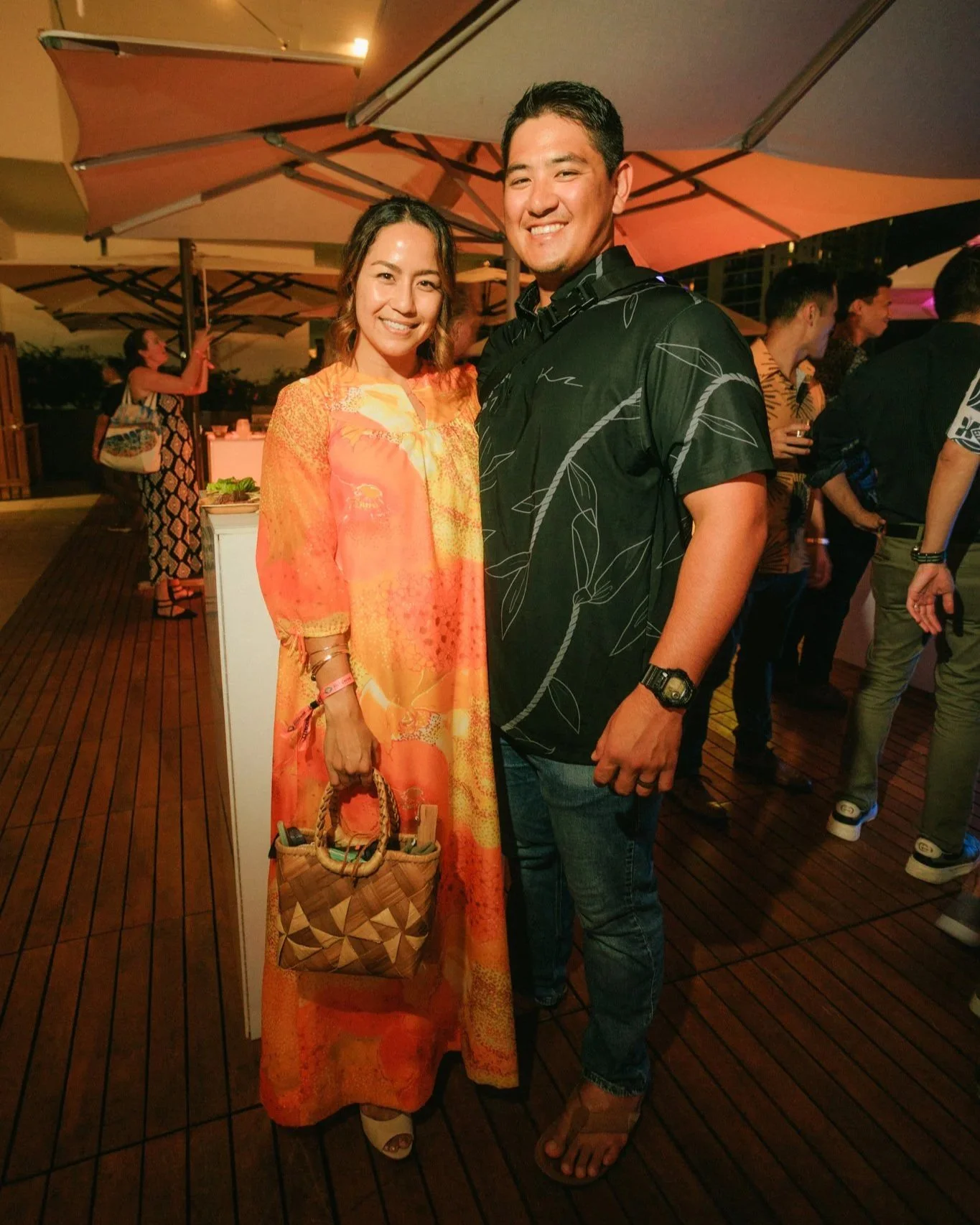 A man and woman smiling at an outdoor event, standing on a wooden deck under large umbrellas with other people in the background.