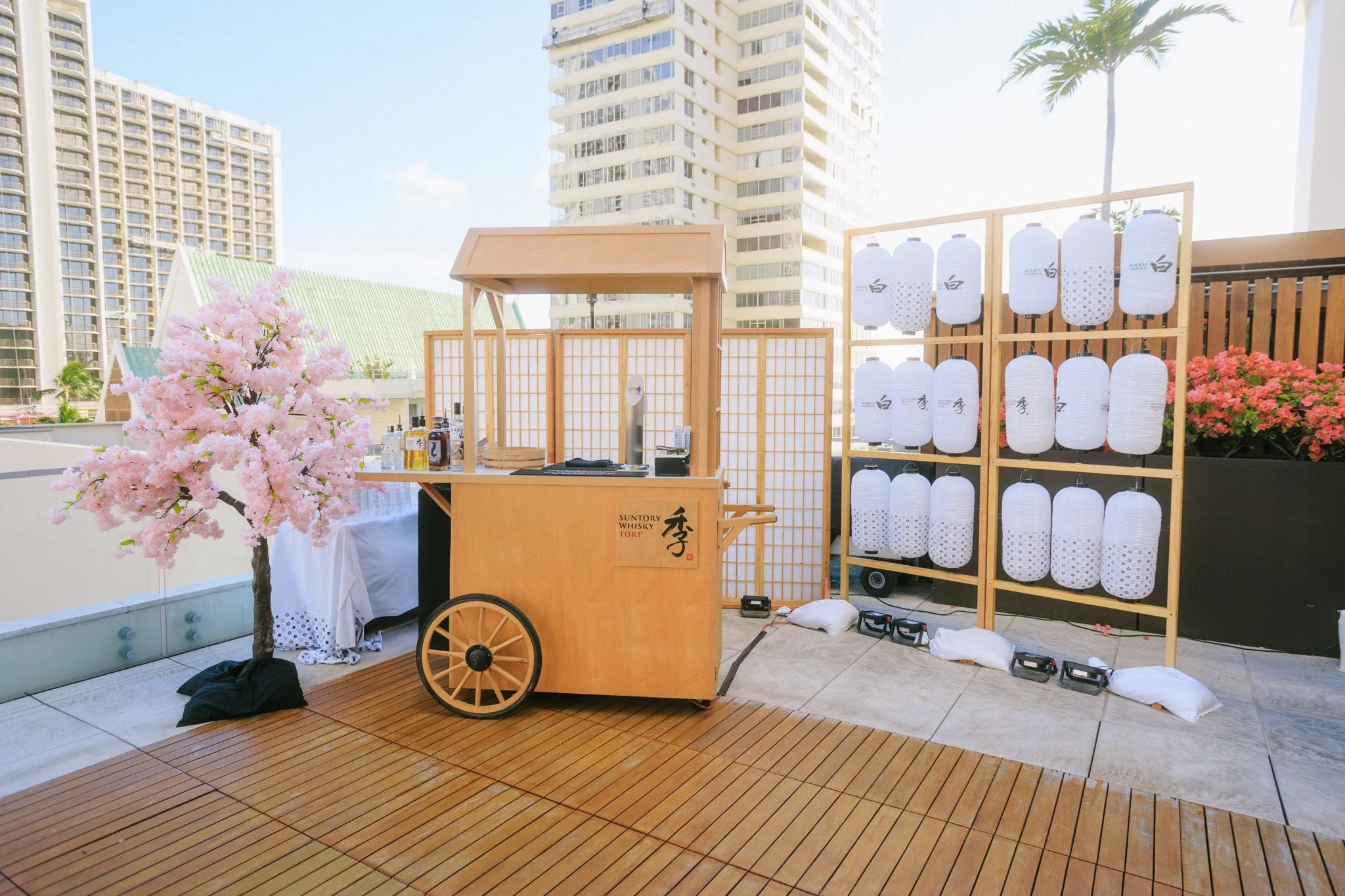 A rooftop setup with a wooden cart, a pink artificial cherry blossom tree, white lanterns, and potted pink flowers, set against tall buildings and a blue sky.