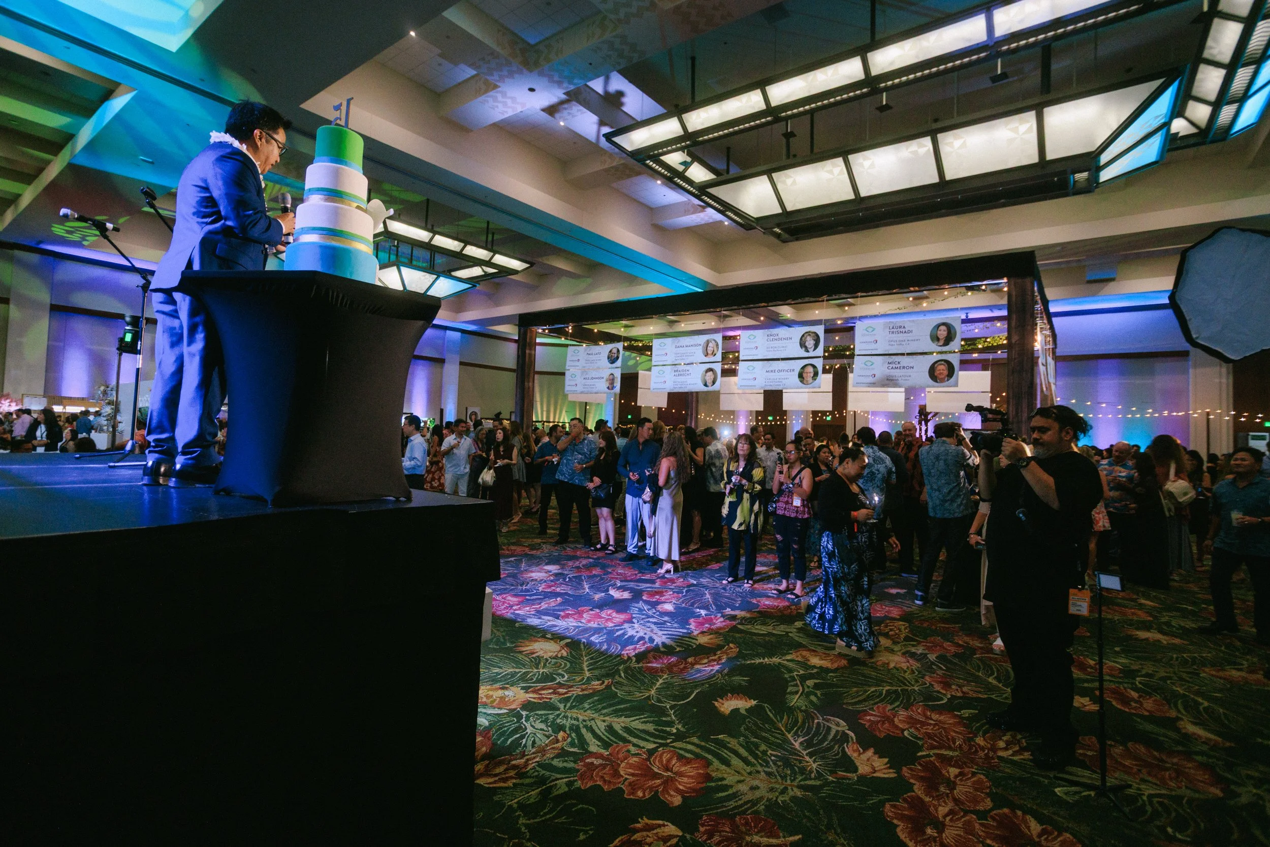A man in a suit stands on a stage next to a large, multi-tiered cake at a crowded conference or event, with attendees mingling and large screens displaying names and photos in the background.