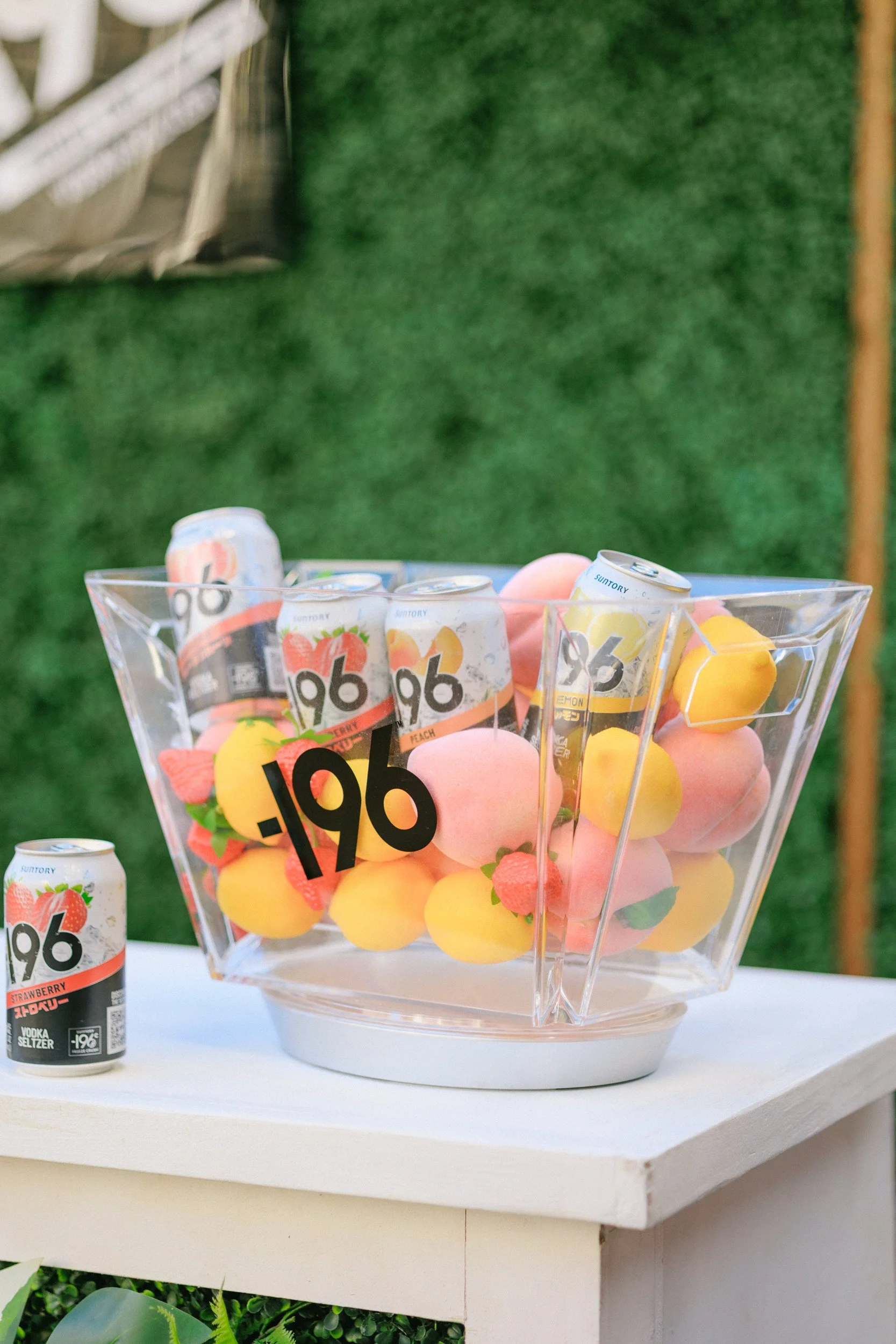 A clear plastic container filled with pink, yellow, and peach-colored round objects resembling foam or plastic balls, with some small artificial strawberries and green leaves added for decoration. The container has a large black label reading '.196' 