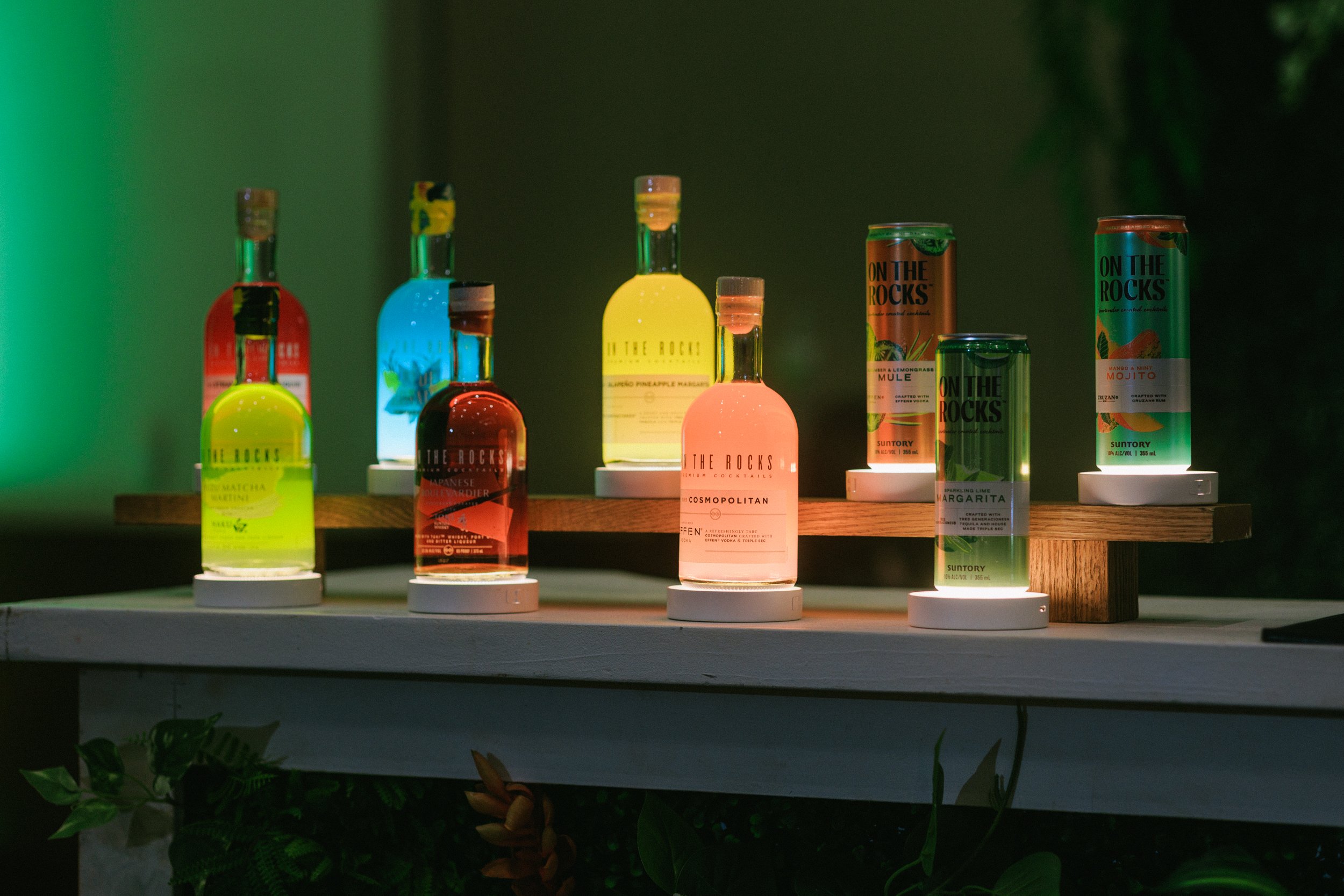 Assorted colorful canned and bottled cocktails displayed on a wooden and white surface with a dark background.