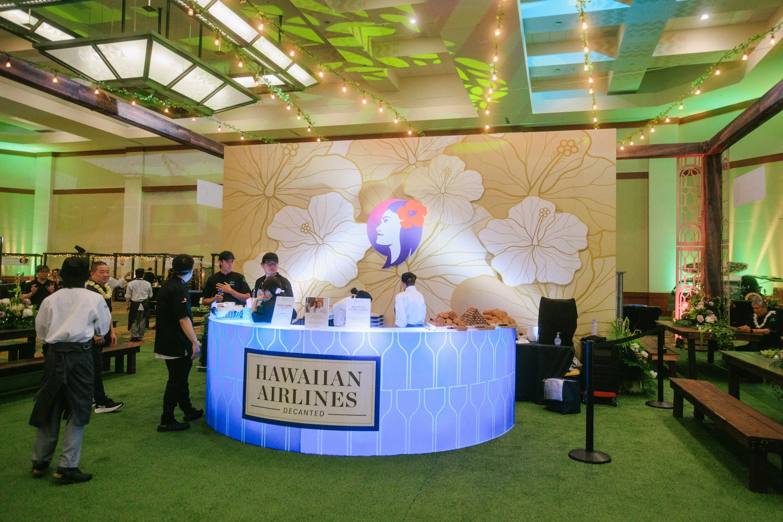 Event featuring Hawaiian Airlines with a luau theme, decorated with large floral wall art, green lighting, and a bar area with staff preparing drinks. Guests are standing around, some engaged in conversation, in a vibrant and festive atmosphere.
