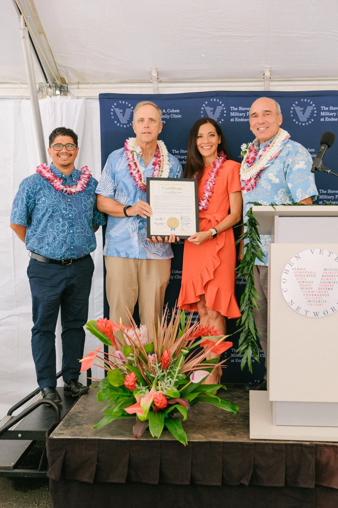 Four people wearing leis, standing on a stage with a certificate and a floral arrangement in front, at a ceremony for the Cohen Veterans Network.