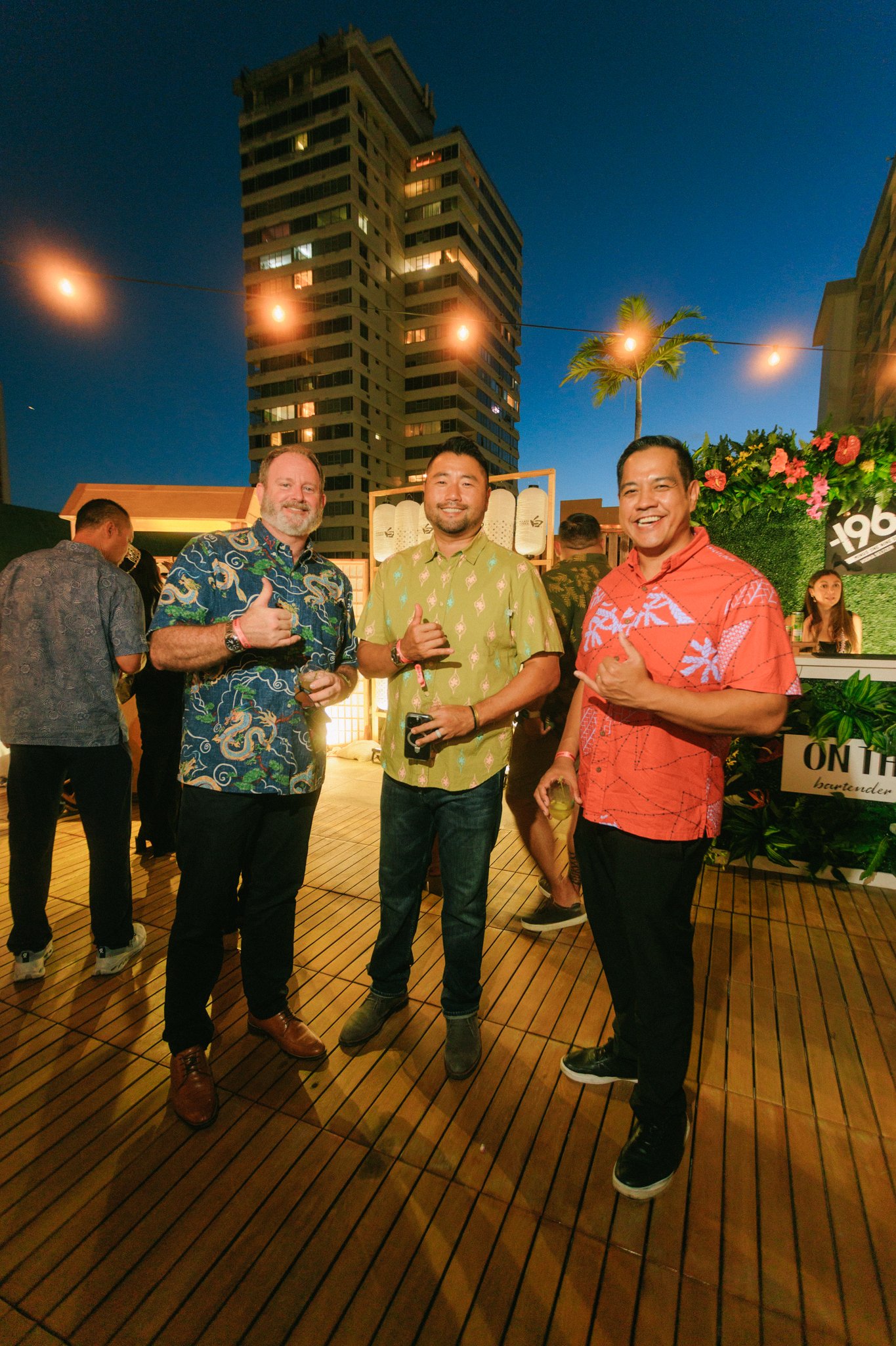 Three men in colorful shirts standing on a wooden deck at an outdoor evening event, smiling and holding drinks, with high-rise buildings, string lights, and a palm tree in the background.