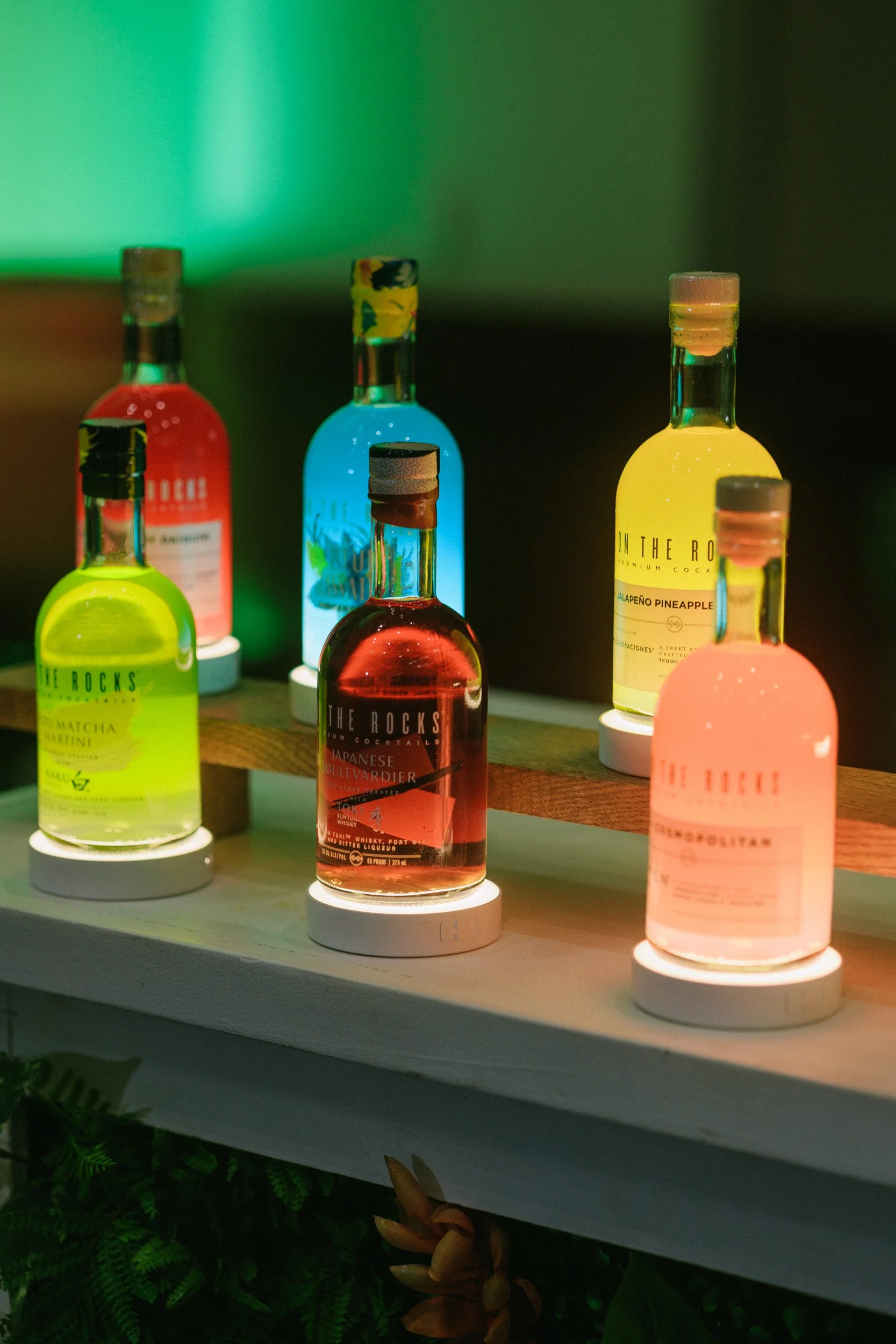Colorful illuminated bottles of cocktails on a wooden shelf with plant decorations below.