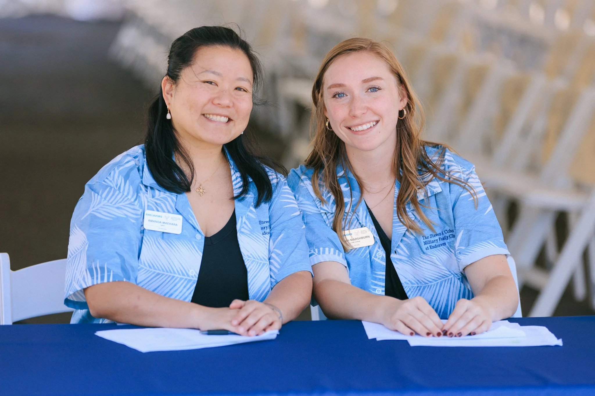 Two women smiling and sitting at a table, wearing blue patterned shirts with name tags, in an indoor setting with white chairs in the background.