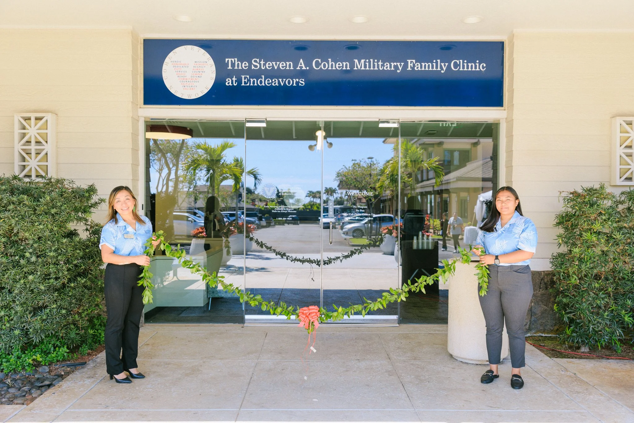 Two women standing outside the entrance of The Steven A. Cohen Military Family Clinic at Endeavors, holding a decorative green leaf garland with a red bow, in front of glass doors and a blue sign.