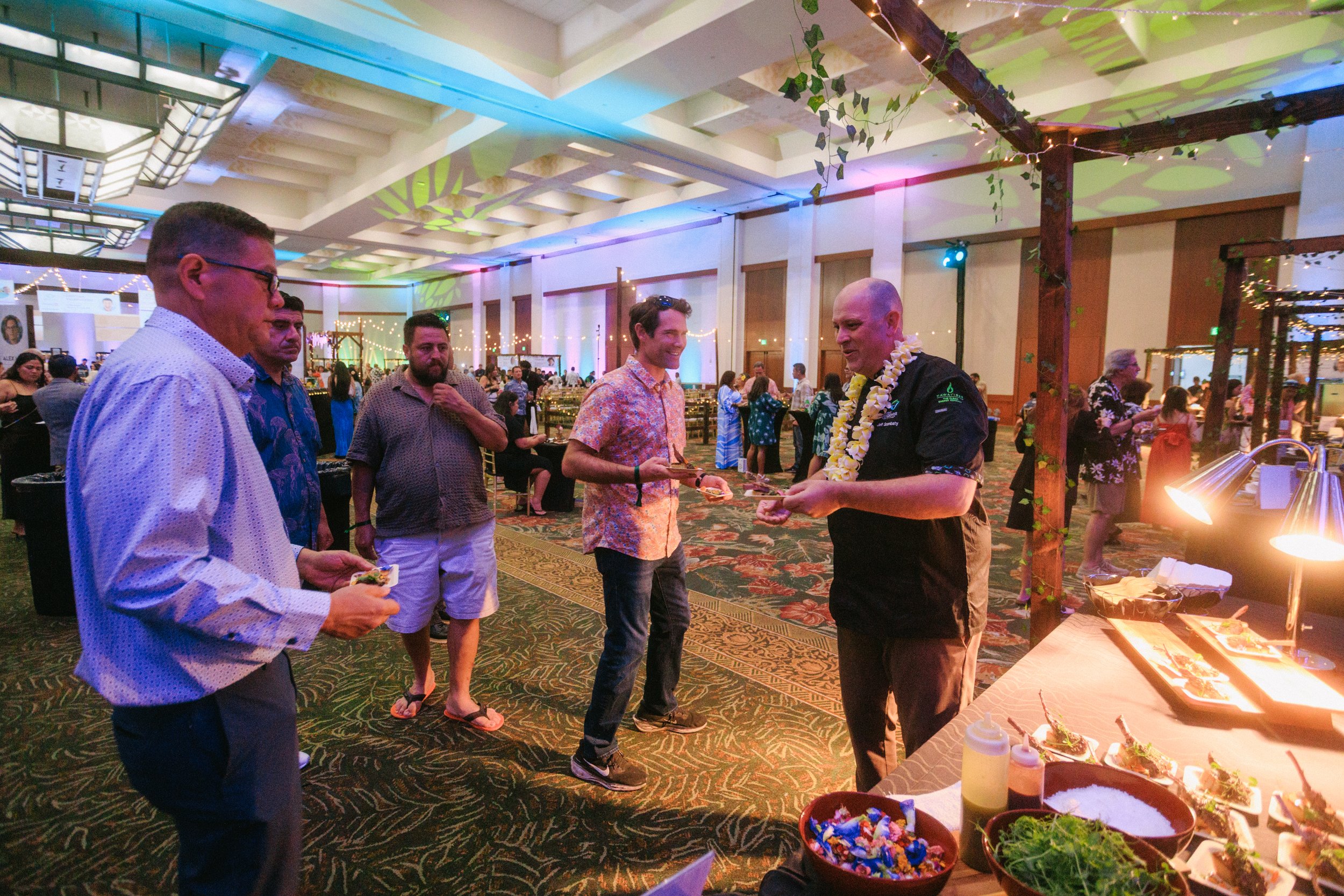 People at a Hawaiian-themed party with food and decorations, including a man wearing a lei, in an event hall with colorful lighting.