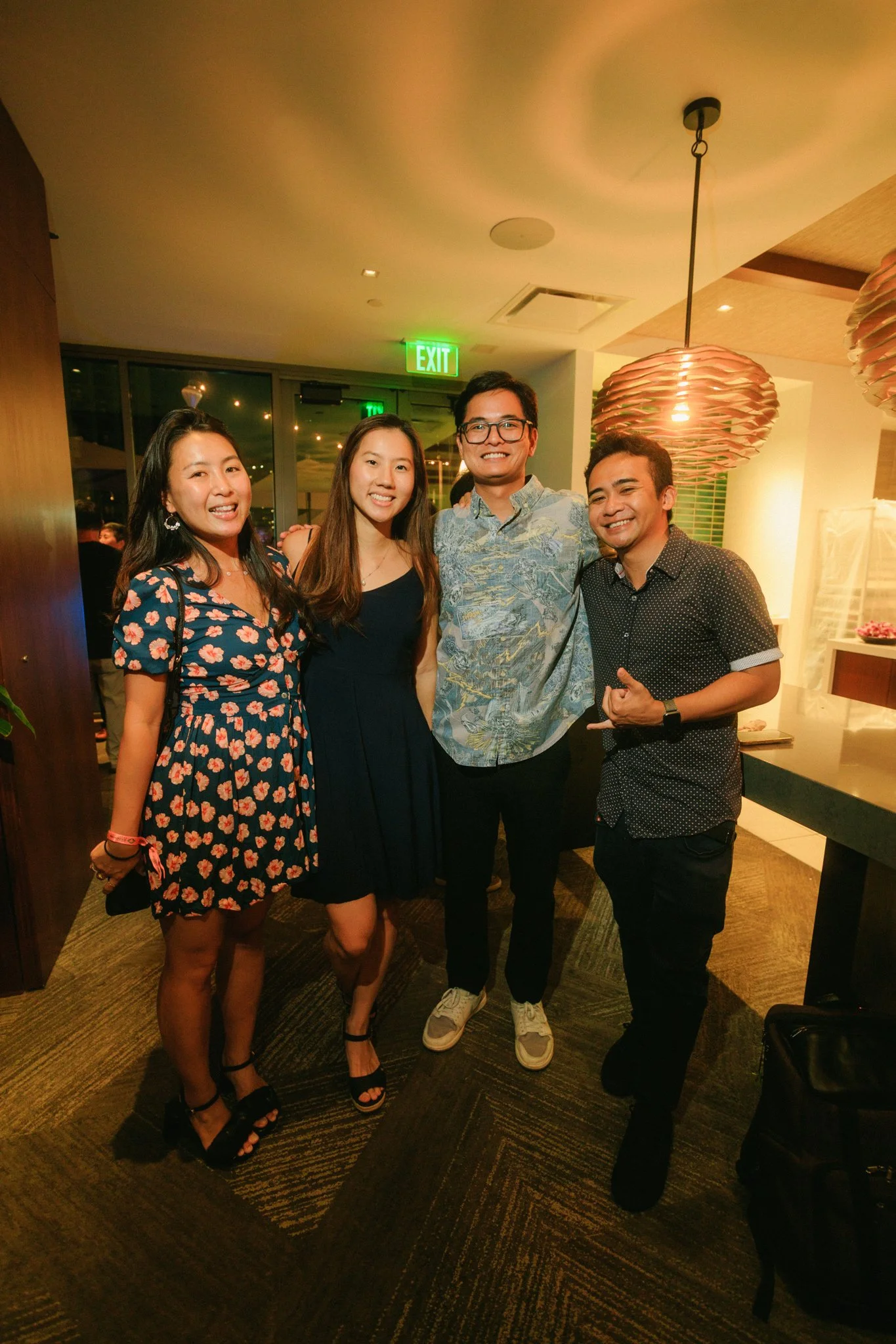 Four young adults smiling and posing together at a social event in an indoor setting with warm lighting.