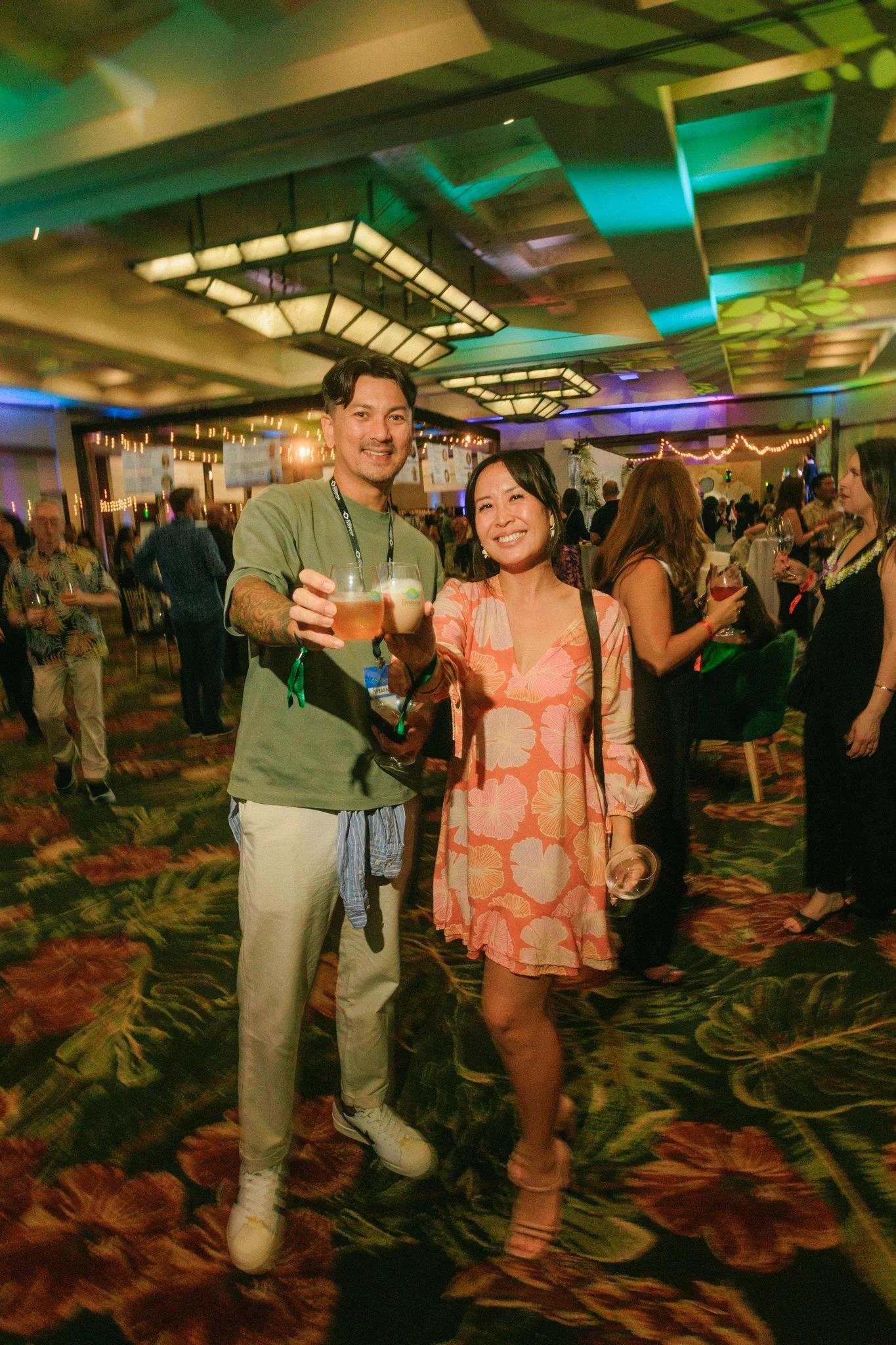 Two people at a social event holding drinks and smiling at the camera, with other guests in the background under colorful lighting.