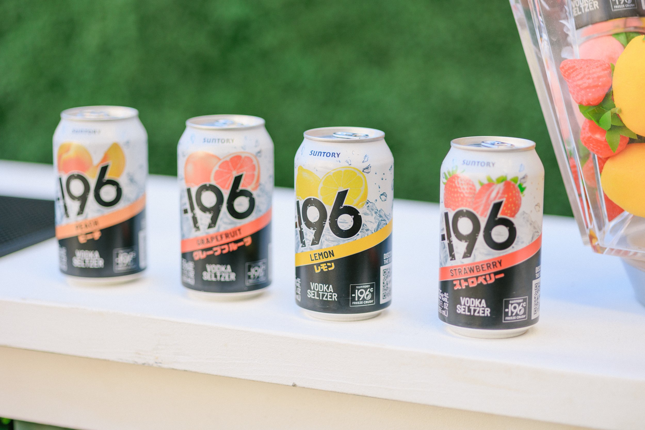 Four cans of 196 vodka seltzer flavors on a white surface with a green background, including peach, grapefruit, lemon, and strawberry.