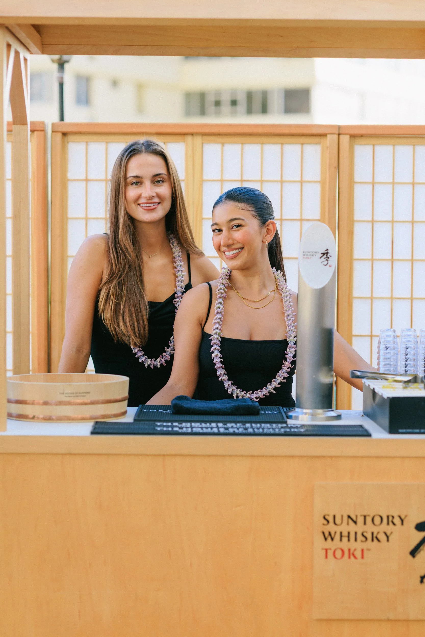 Two women standing behind a wooden beverage cart with leis around their necks, smiling. The cart has a sign that reads 'Suntory Whisky Toki' and is set outdoors, with a wooden fence and buildings in the background.