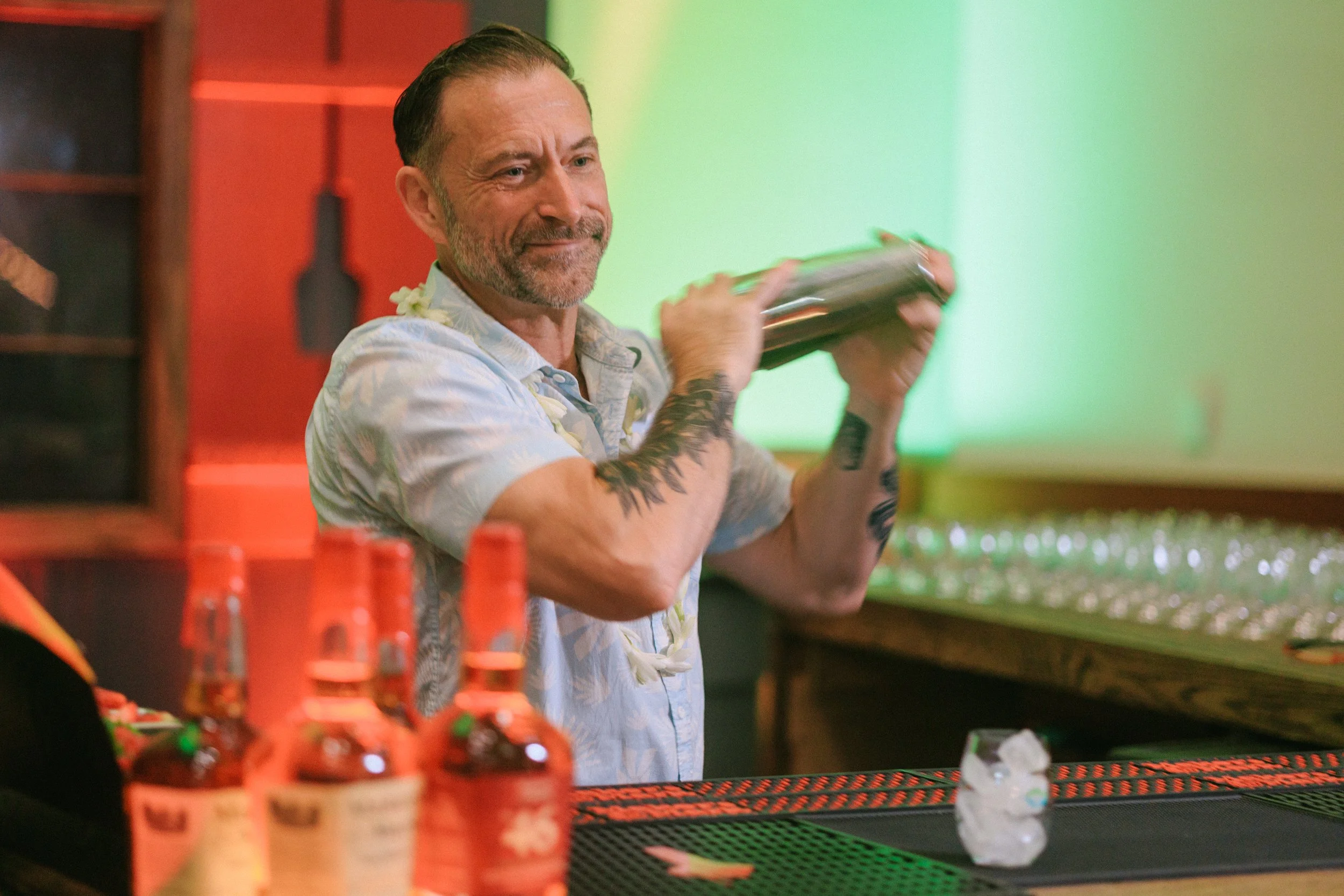 A man with tattoos and a short beard, wearing a light-colored floral shirt, is behind a bar, shaking a cocktail shaker with a smile. In front of him on the counter are several bottles of hot sauce or similar condiment, and a small clear glass with ic