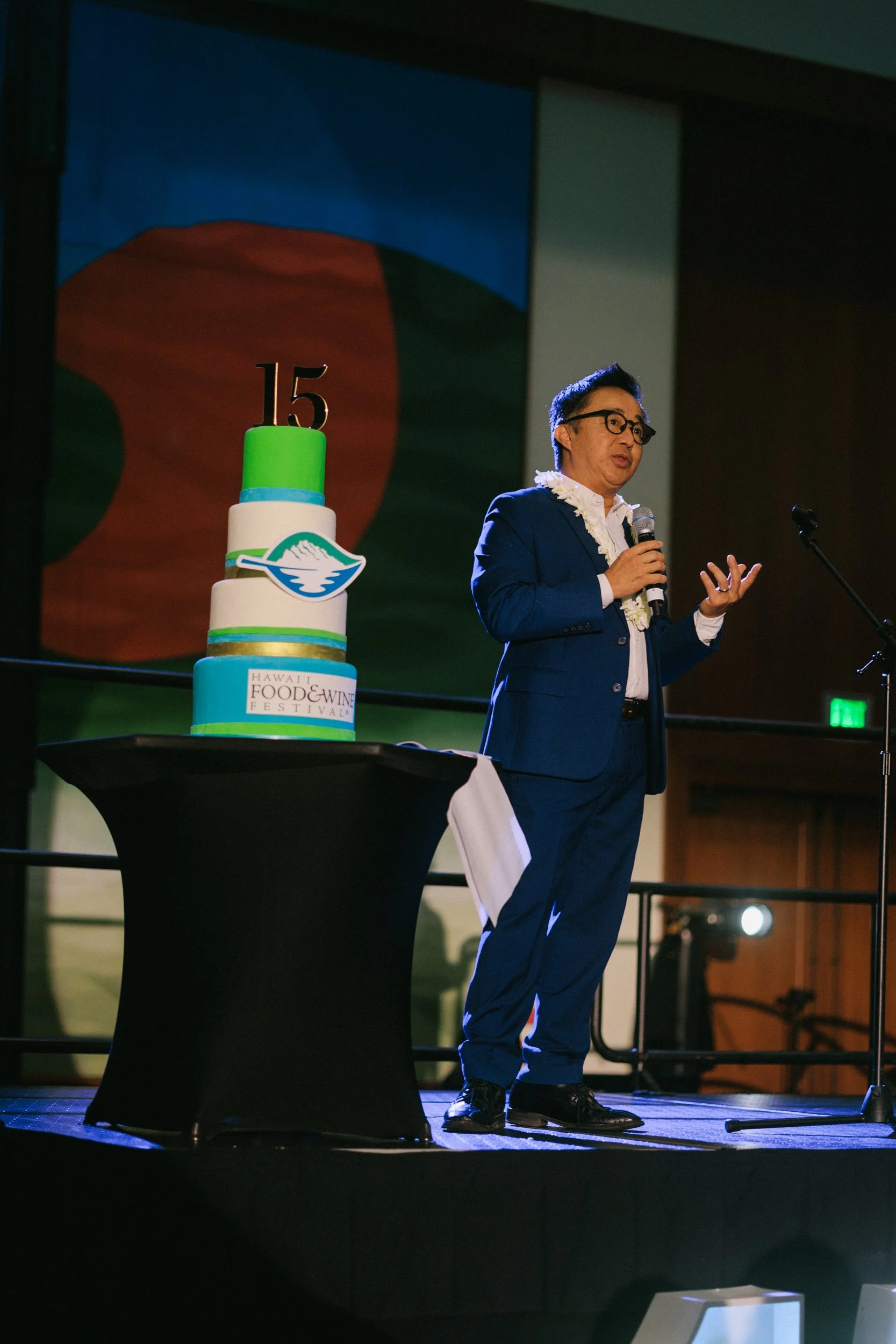 A man in a blue suit and glasses speaking into a microphone on stage next to a four-tiered cake with a green and white color theme, featuring a '15' topper and the Hawaii Food & Wine Festival logo, in a venue with artistic background.