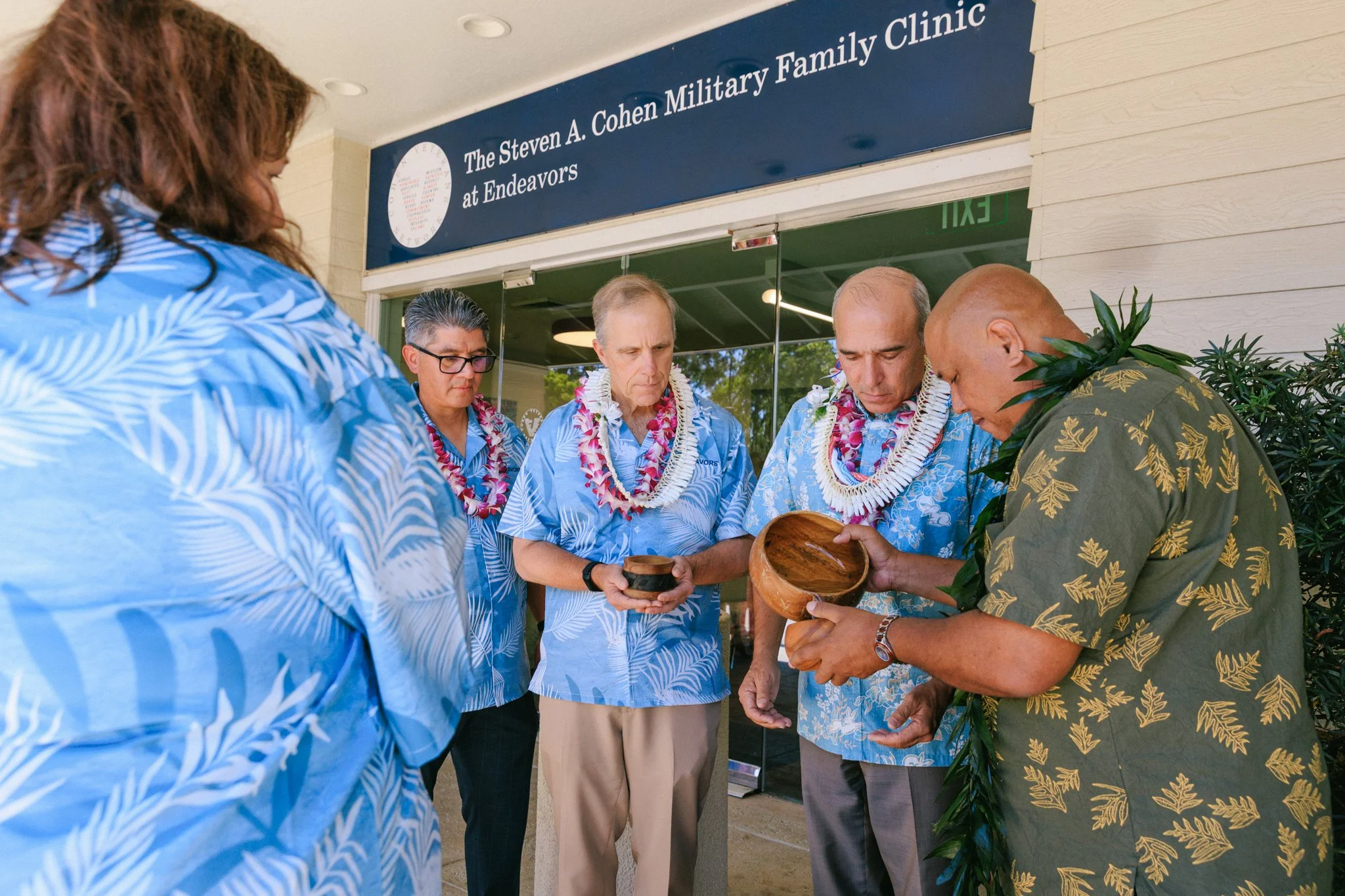 A group of men and women in Hawaiian shirts and leis gathered outside The Steven A. Cohen Military Family Clinic at Endeavors, participating in a ceremonial activity involving wooden bowls.