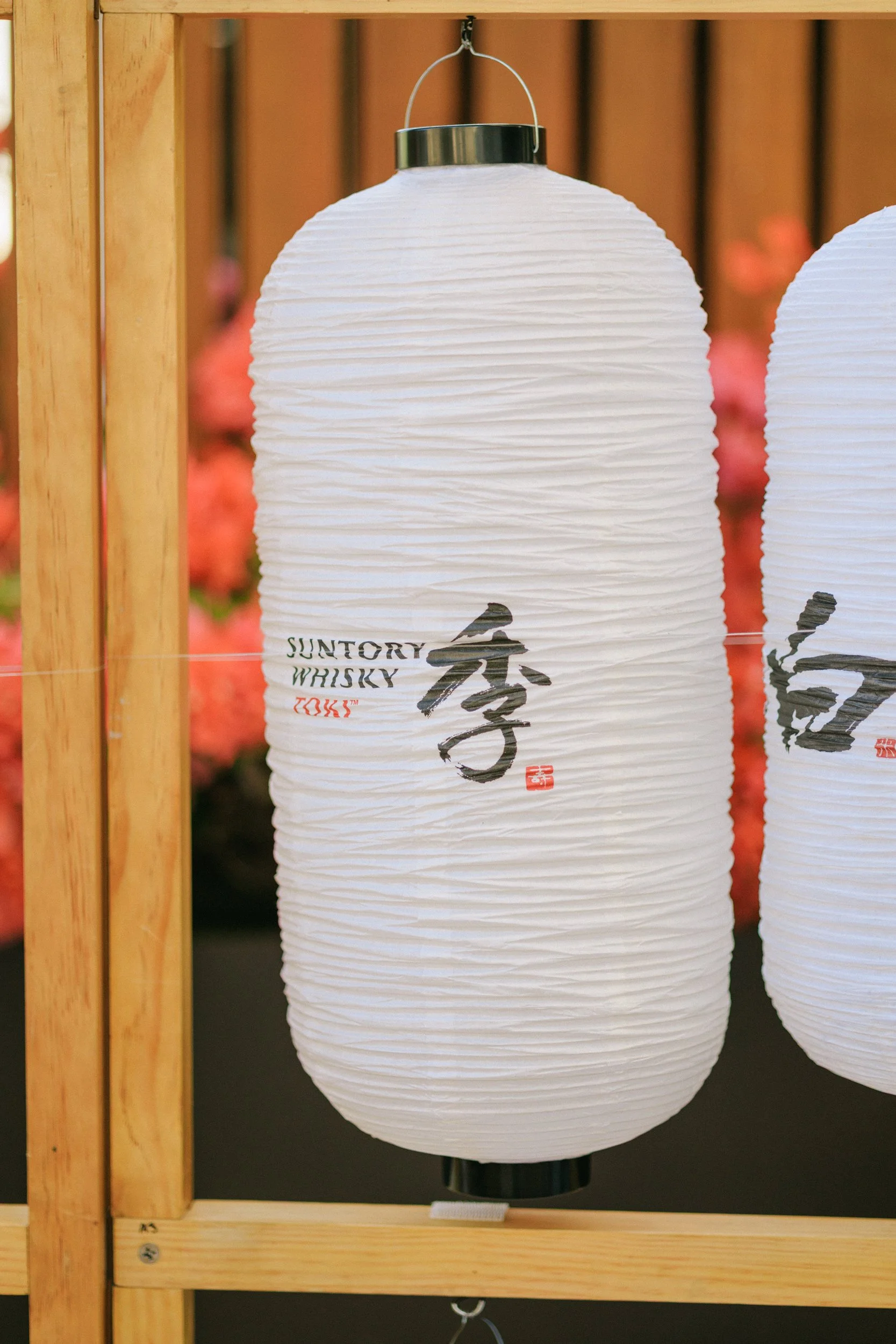 White paper lantern with black and red Japanese characters, hanging on a wooden frame, with blurred pink flowers in the background.