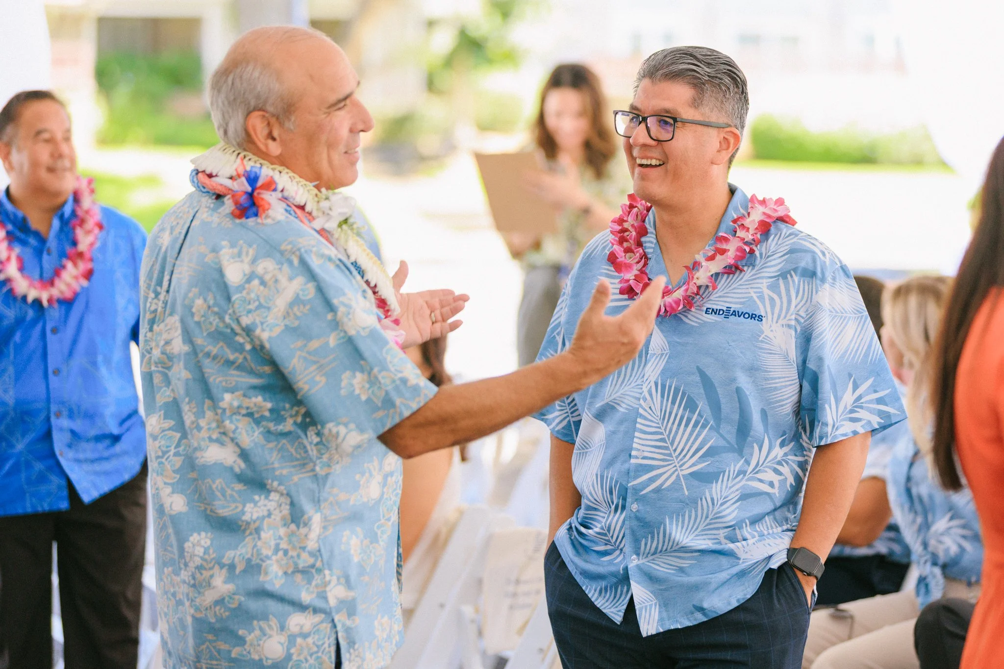 People at a Hawaiian-themed outdoor party wearing flower leis and tropical shirts, engaging in conversation and smiling.