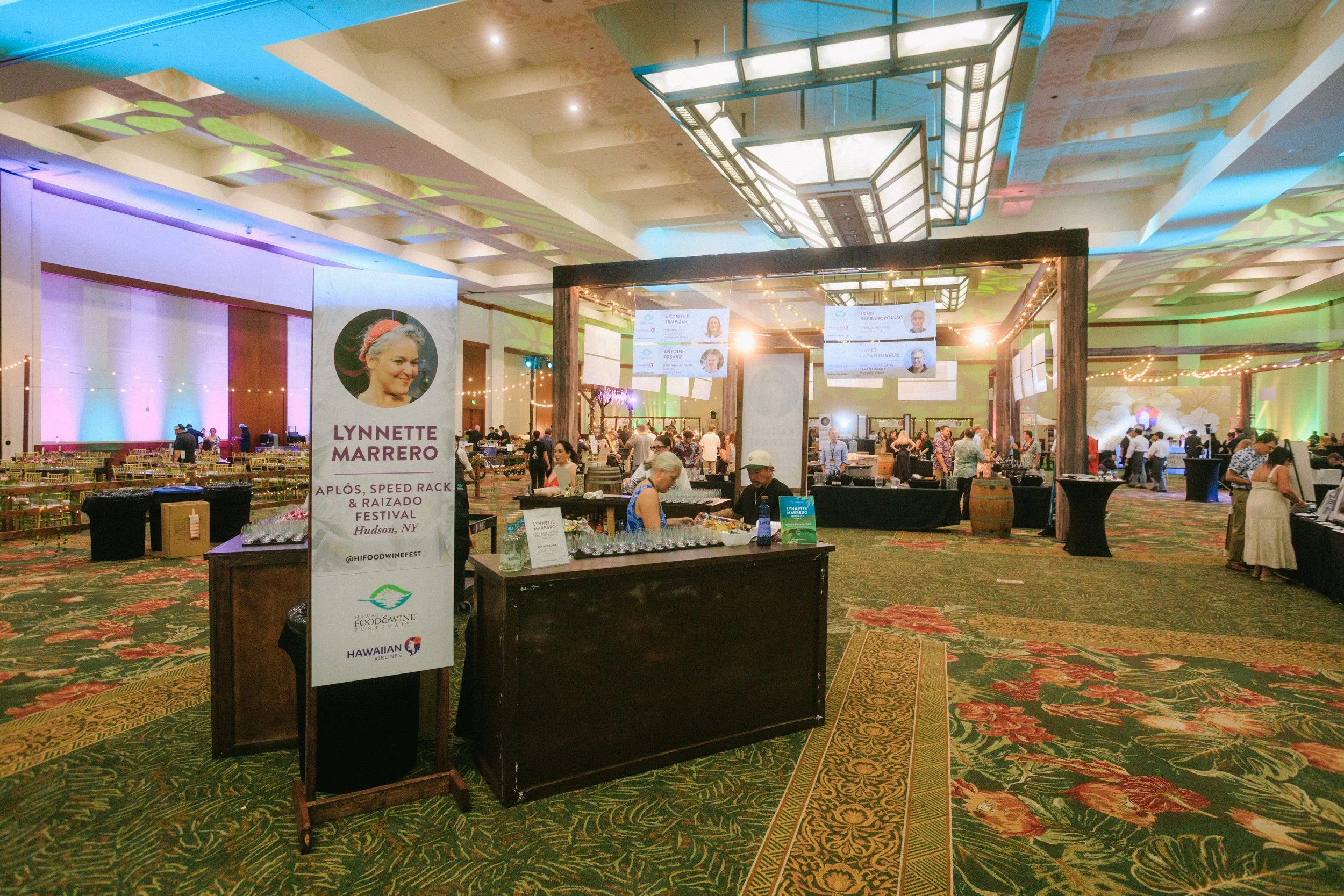 Indoor event with tables, screens, and people, featuring a sign for Lynette Marrero at the Ap l's, Speed Rack & Raizado Festival in Hudson, NY, decorated with string lights and colorful lighting.