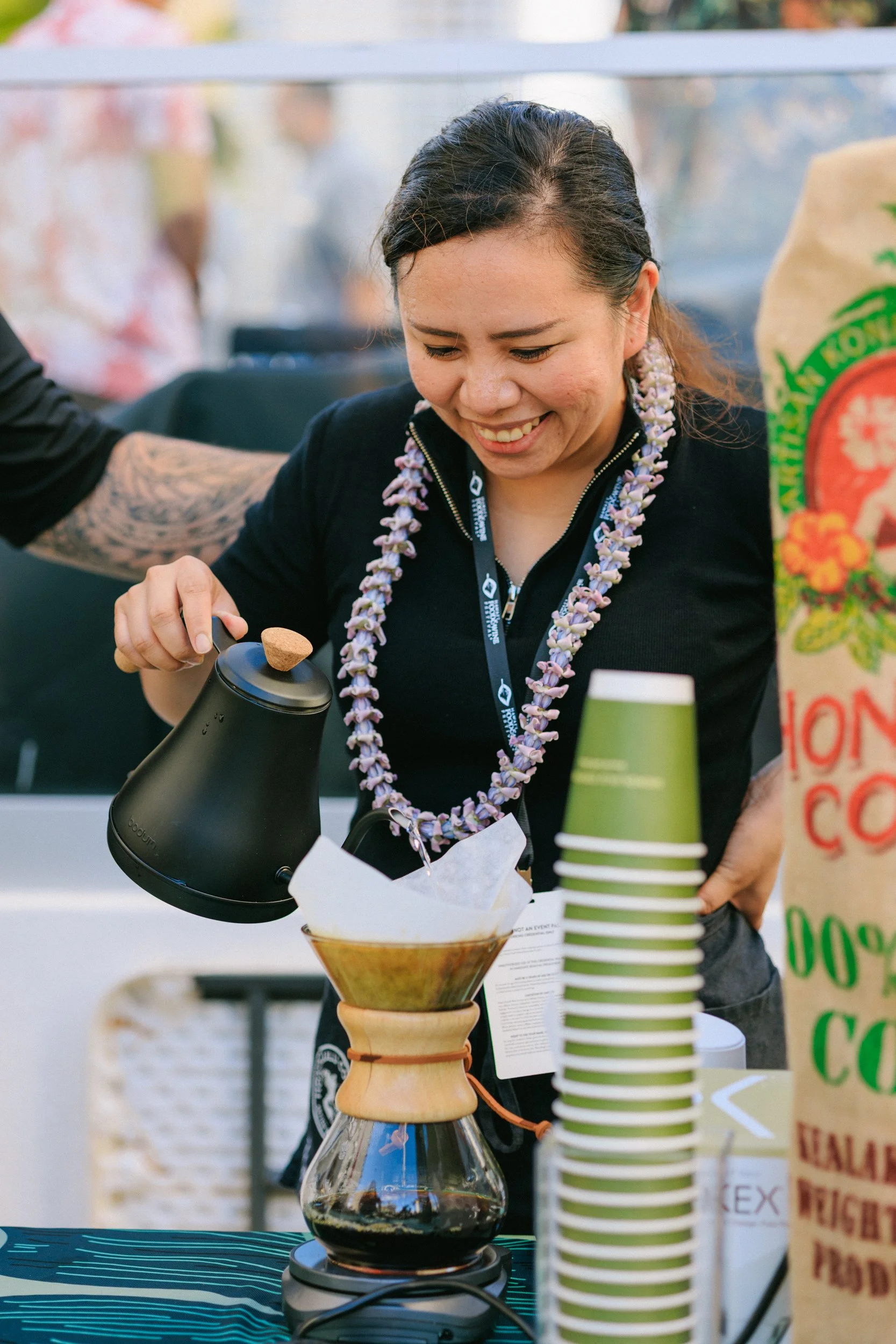 A woman with a purple floral lei around her neck is pouring water from a black gooseneck kettle into a coffee brewing device with a paper filter. She is smiling and wearing a black jacket, standing at a table with stacks of green paper cups, a coffee