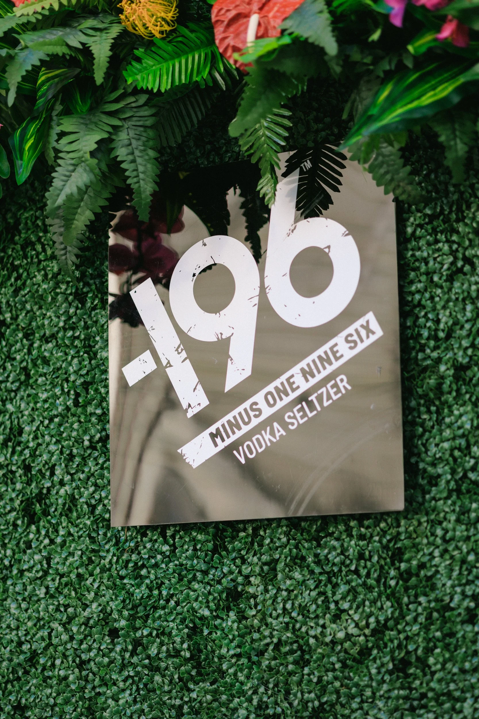Sign on a shiny metallic surface with large, distressed white numbers reading '190' and smaller text indicating a cocktail, 'Minus One Nine Six Vodka Seltzer,' surrounded by green artificial foliage and flowers.