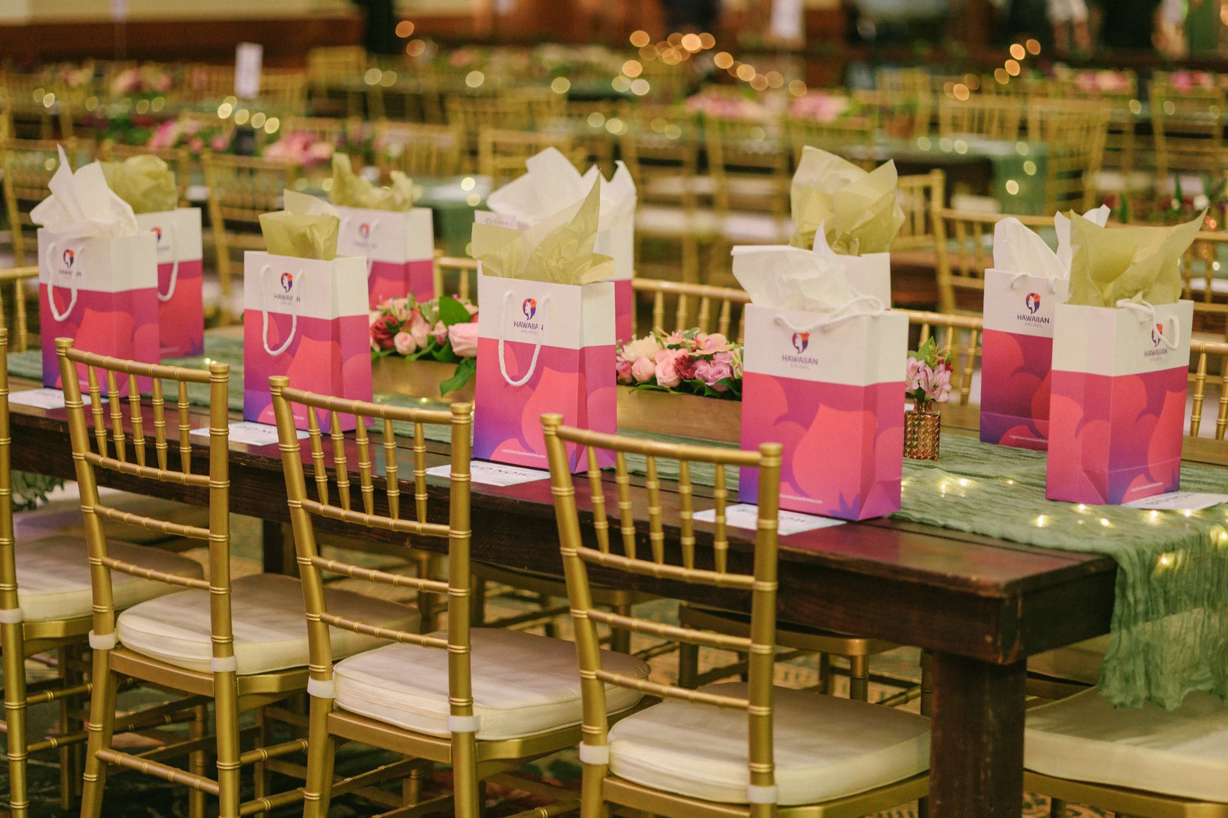 A decorated banquet table set for an event, with gold chairs, pink gift bags with tissue paper, and pink floral centerpieces. The table has a green tablecloth with string lights, and the background shows more chairs and decorations.