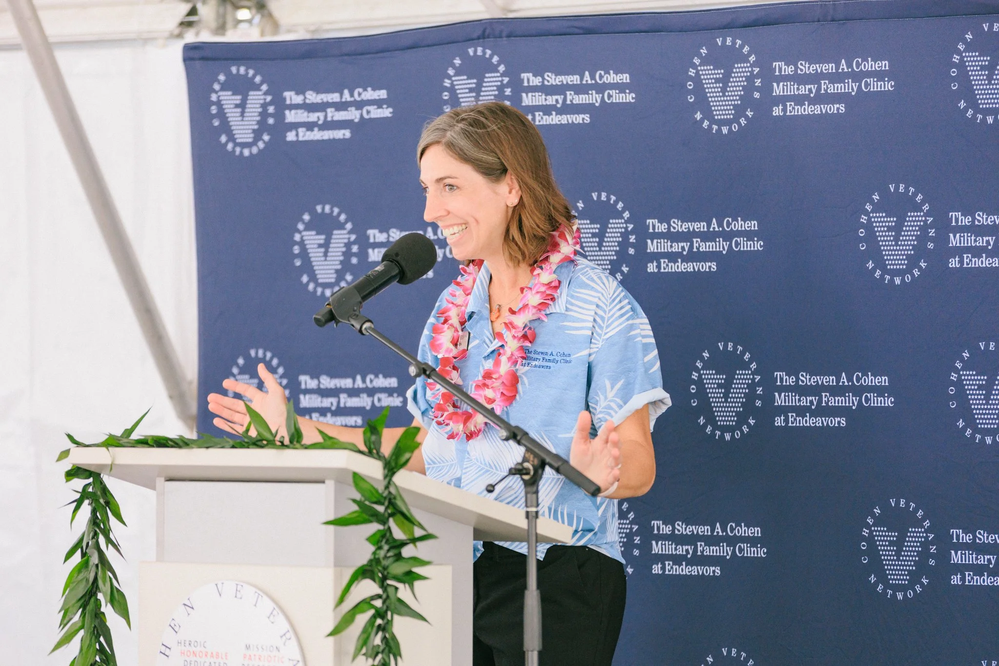 A woman speaking at a podium with a blue backdrop that reads "The Steven A. Cohen Military Family Clinic at Endeavors." She has shoulder-length brown hair, is wearing a blue patterned shirt, a pink lei, and is gesturing with her hands.