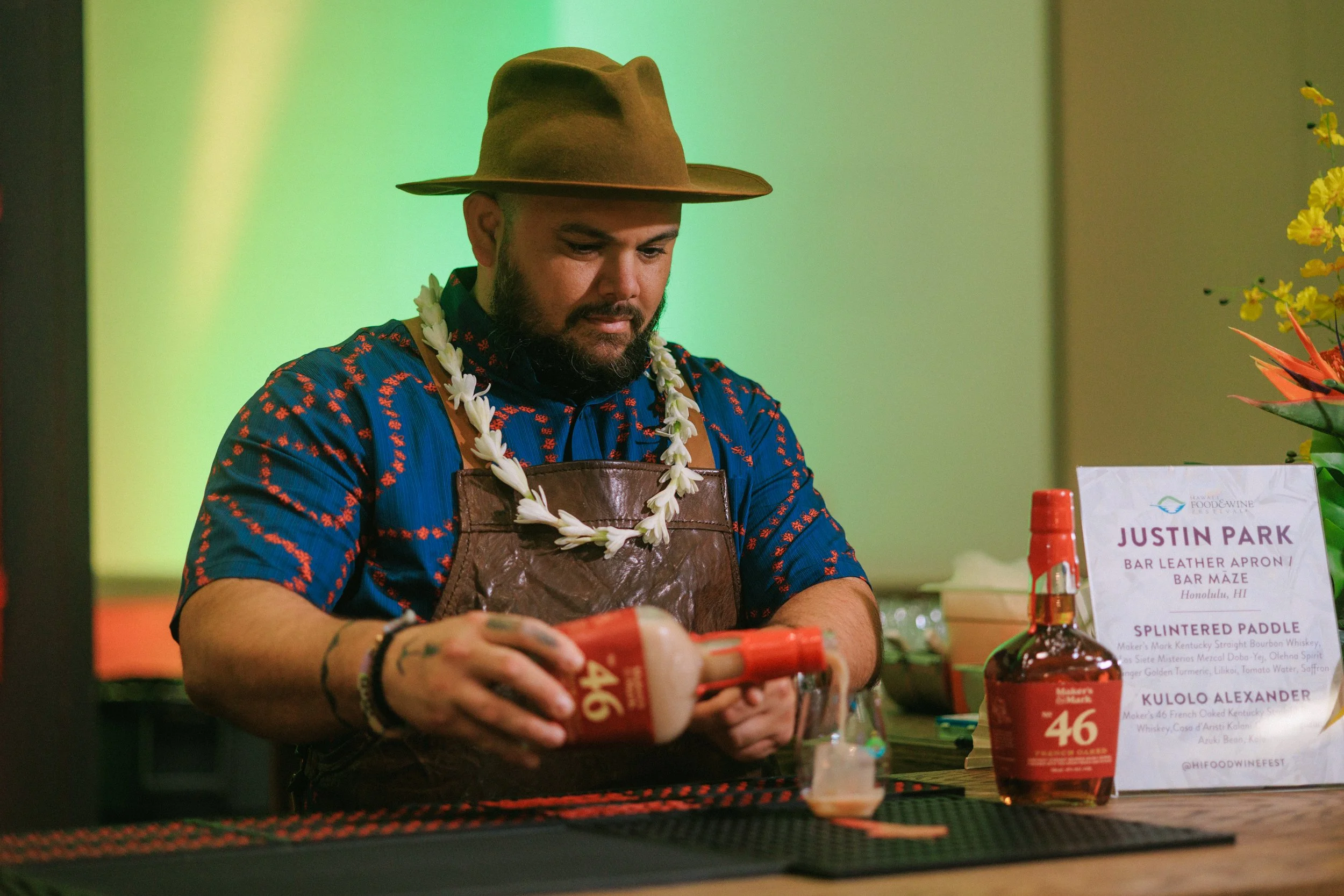 Man wearing a brown hat, a patterned blue shirt, a leather apron, creating a cocktail behind a bar with a sign reading Justin Park and a bottle of Maker's Mark bourbon.