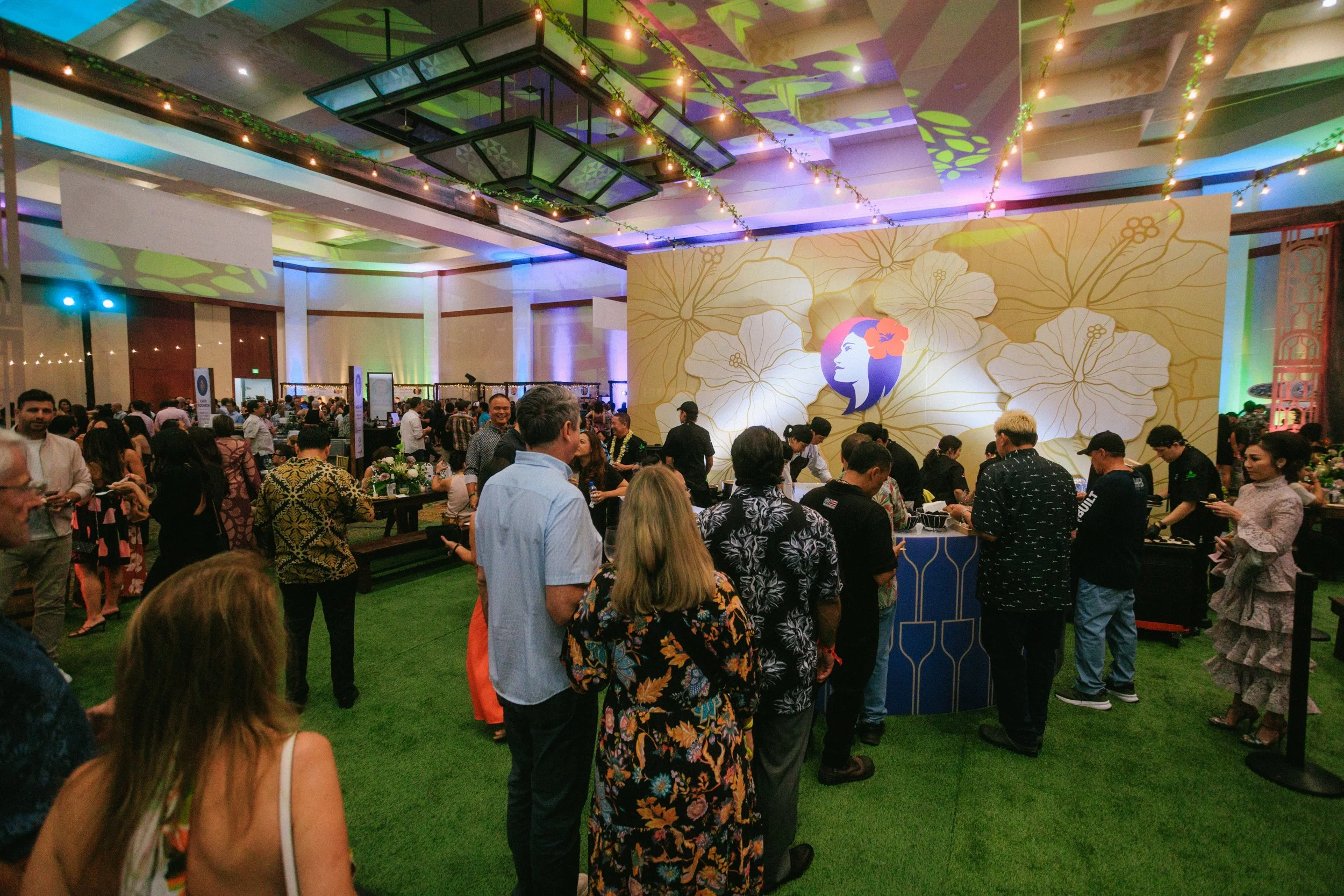 A bustling indoor event with many people gathered around a decorated booth featuring a large floral wall with a silhouette of a woman's face with flowers in her hair. The venue has string lights, colorful lighting, and a green carpeted floor.
