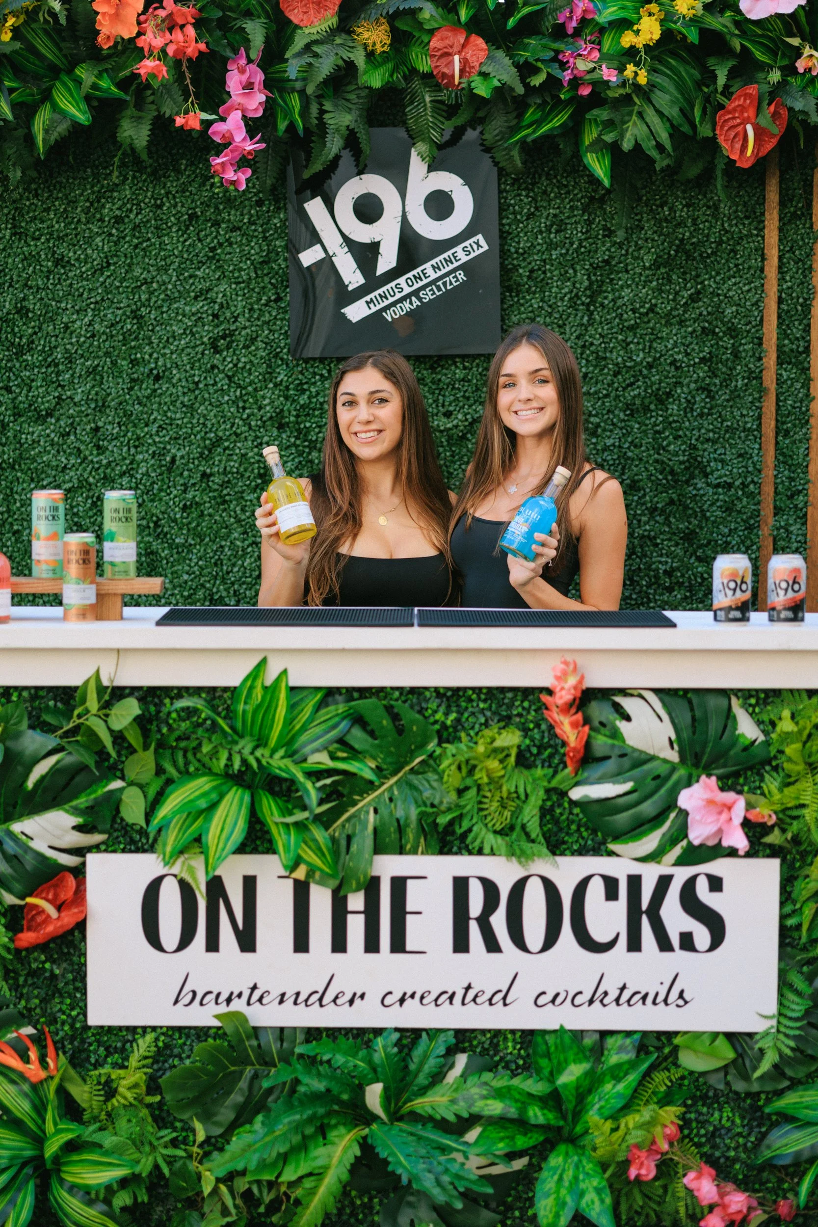 Two women behind a bar with a green leafy background and floral decoration, holding colorful bottles of cocktails, with signs reading 'Minus One Nine Six Vodka Seltzer' and 'On the Rocks bartender created cocktails'.