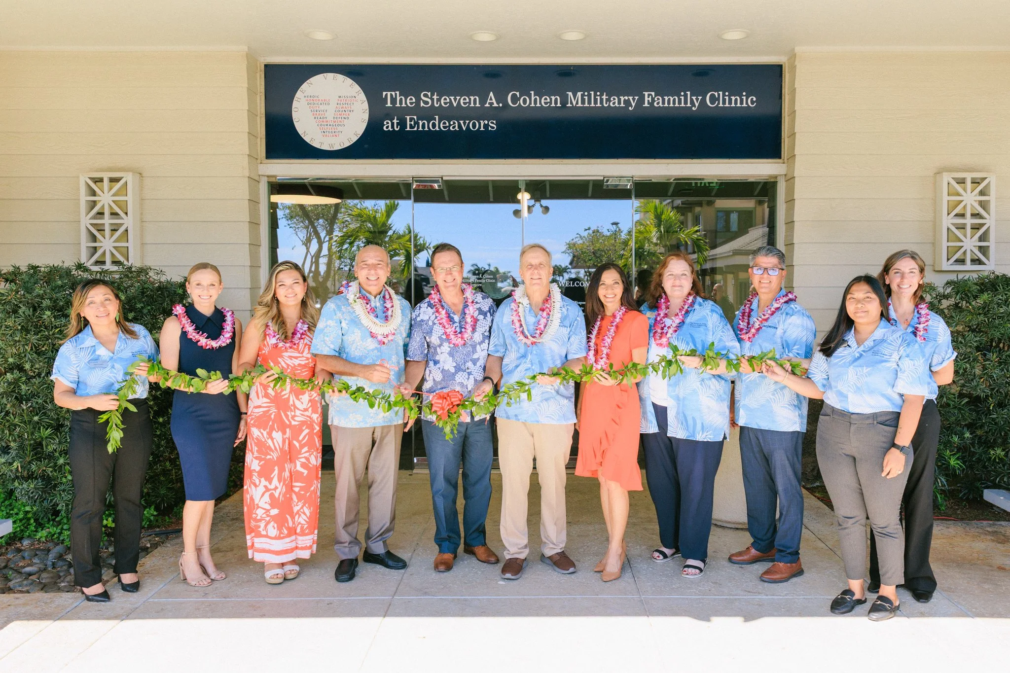 Group of people standing in front of the Steven A. Cohen Military Family Clinic at Endeavors, holding a garland, participating in a ribbon-cutting ceremony.